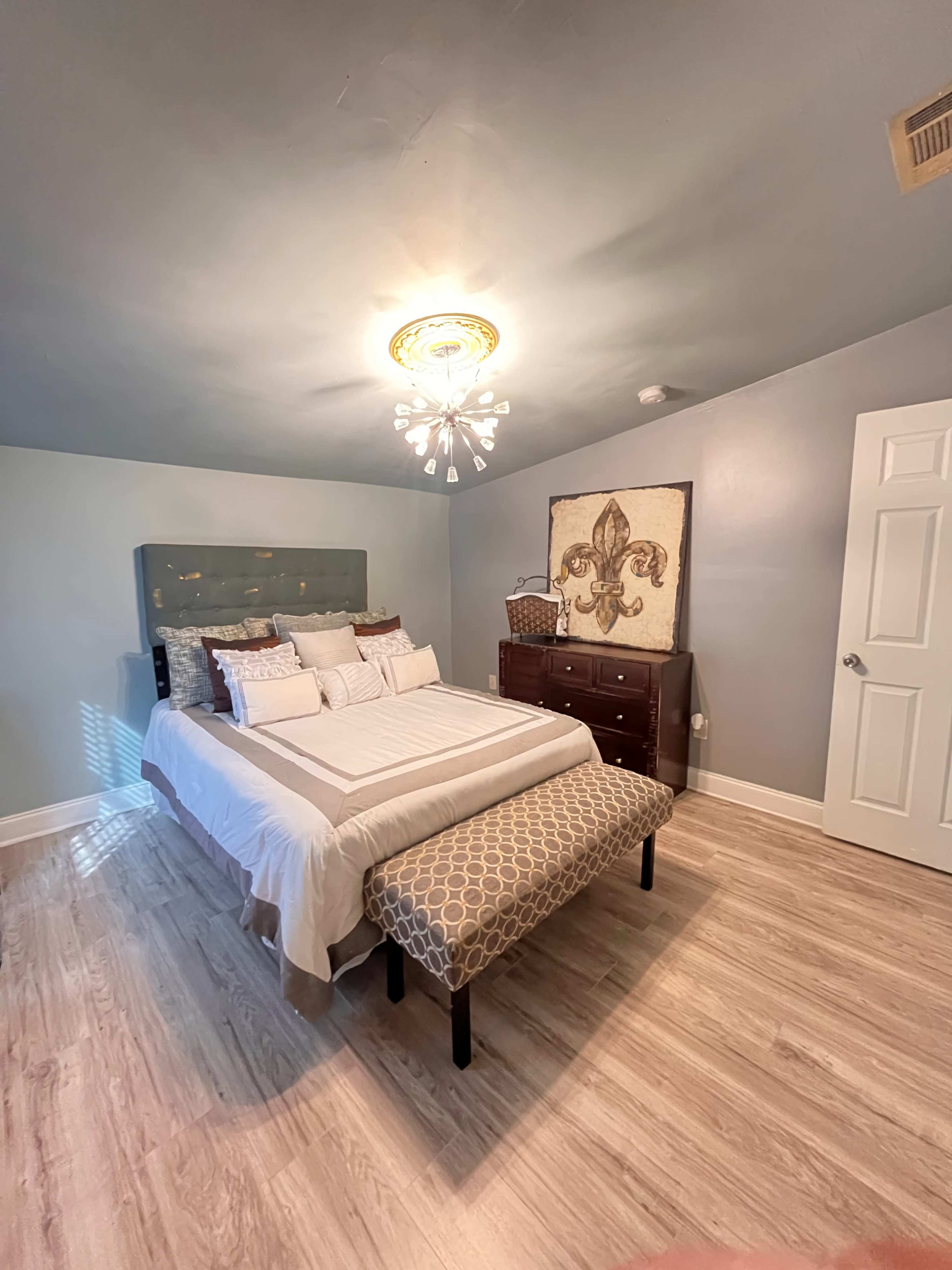 The image shows a well-furnished bedroom featuring a bed with a decorative headboard, a bench at the foot, and a dresser against the wall, with soft lighting from a chandelier.