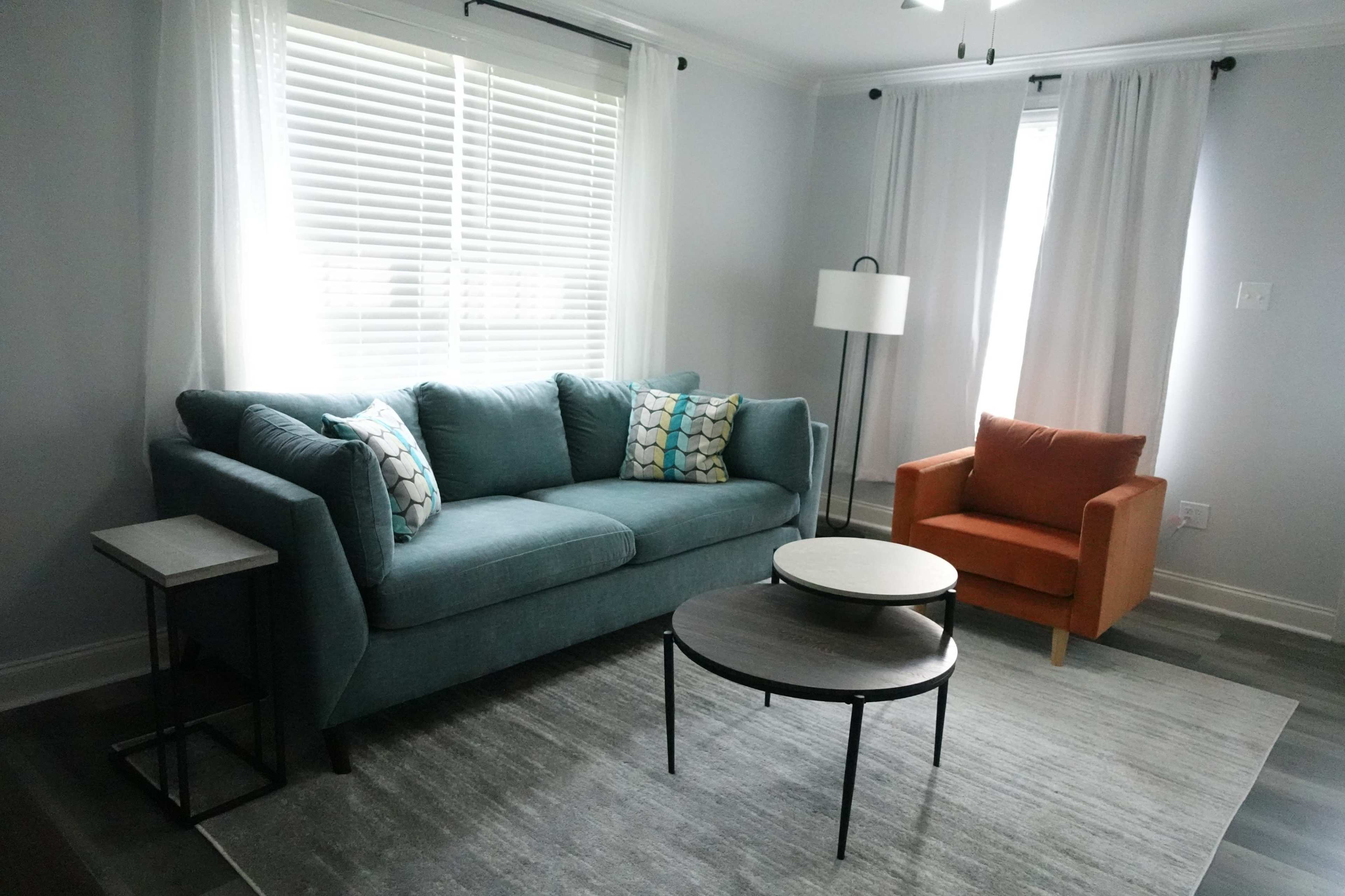 A modern living room with a teal sofa, an orange armchair, a round coffee table, and large windows with white curtains.