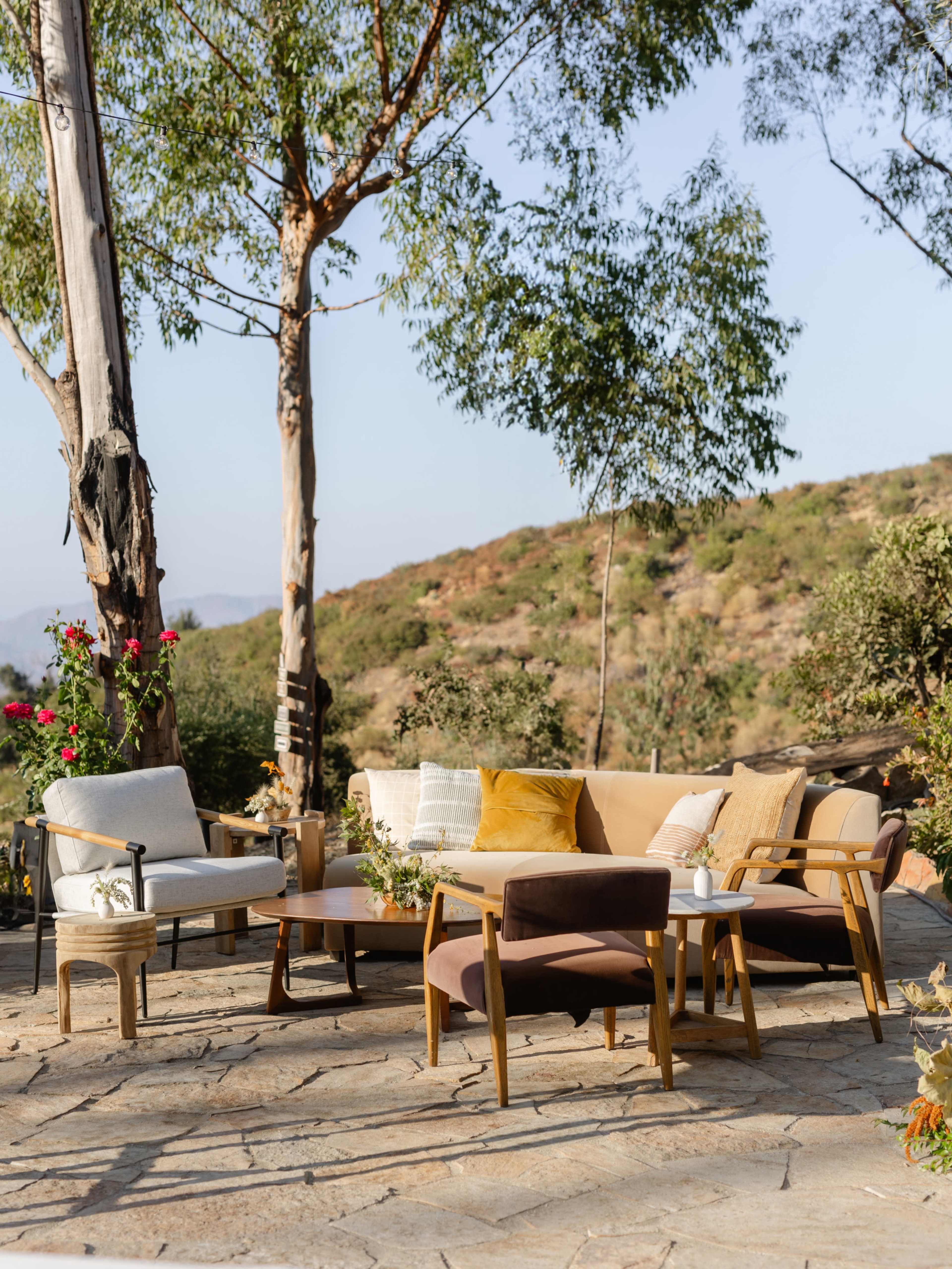 A modern outdoor seating area features a mix of chairs and a sofa arranged around a coffee table on a stone patio, surrounded by trees and hills.