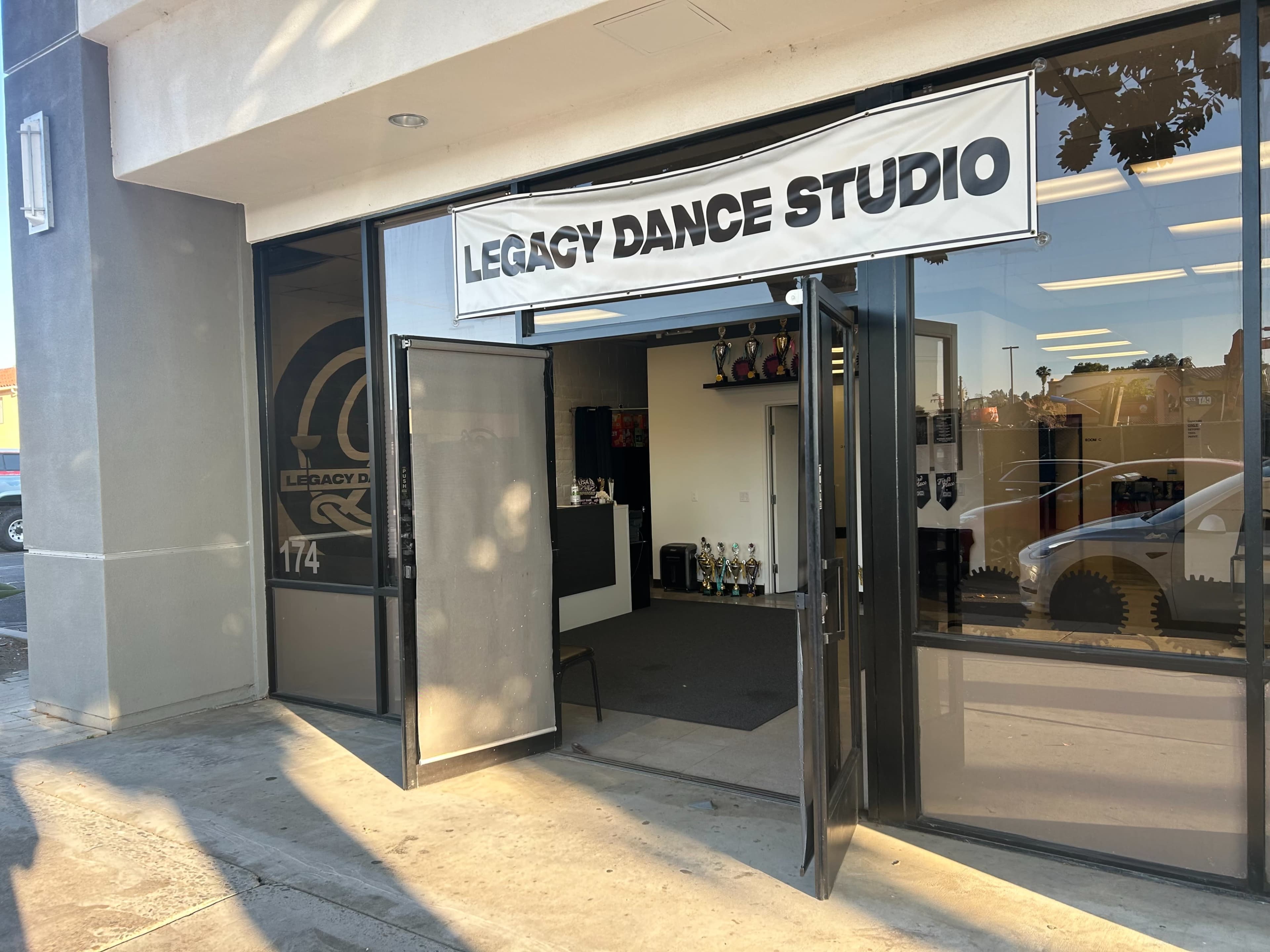 The image shows the entrance of Legacy Dance Studio, featuring a large sign and a display of trophies inside.