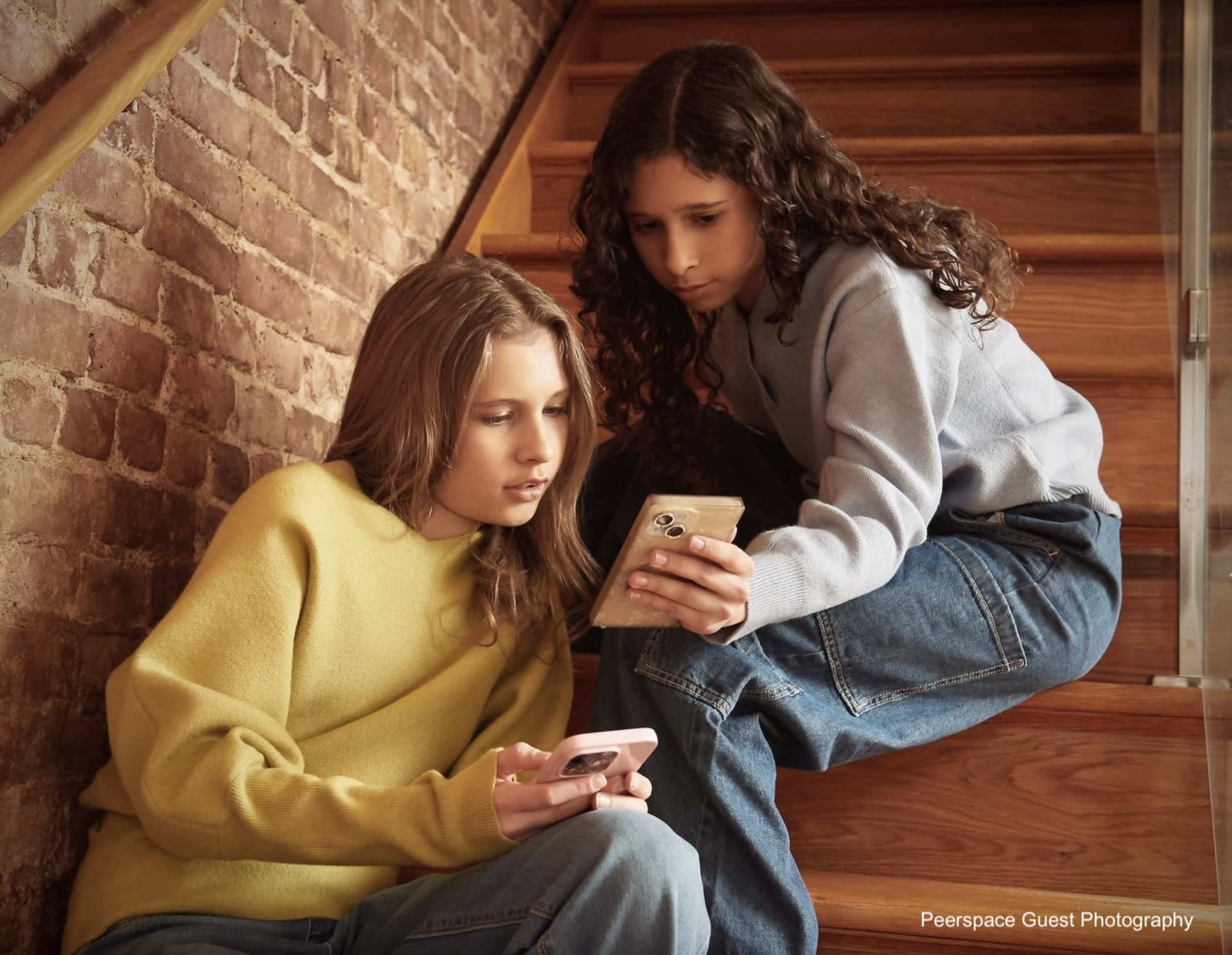 Two girls sit on wooden stairs, each focused on their smartphones.