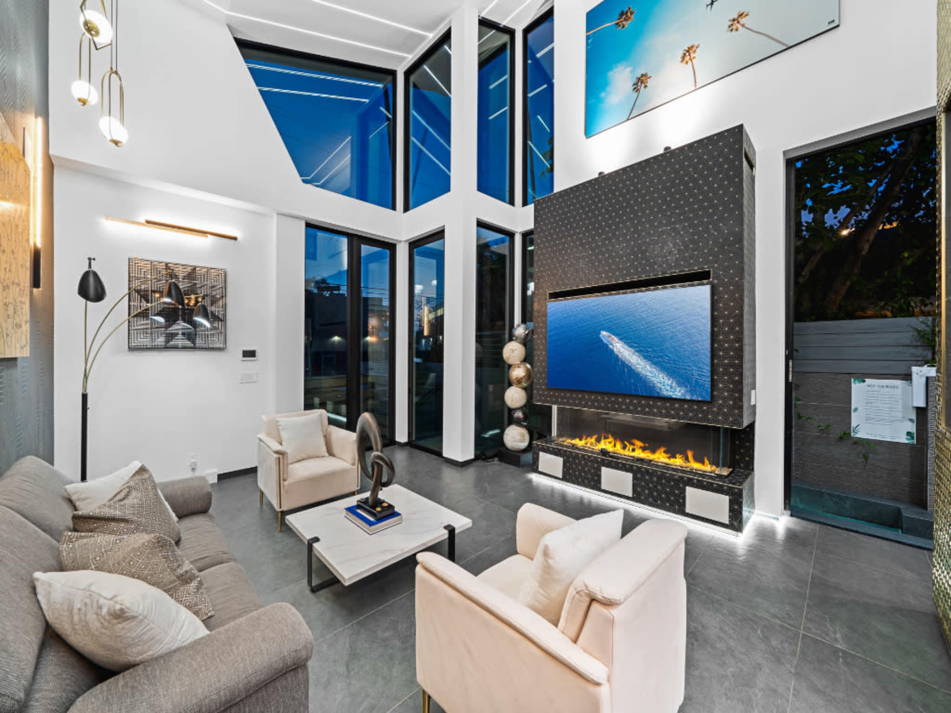 A modern living room features large windows, a sleek fireplace, and a contemporary television, with a sofa and chairs arranged around a coffee table.