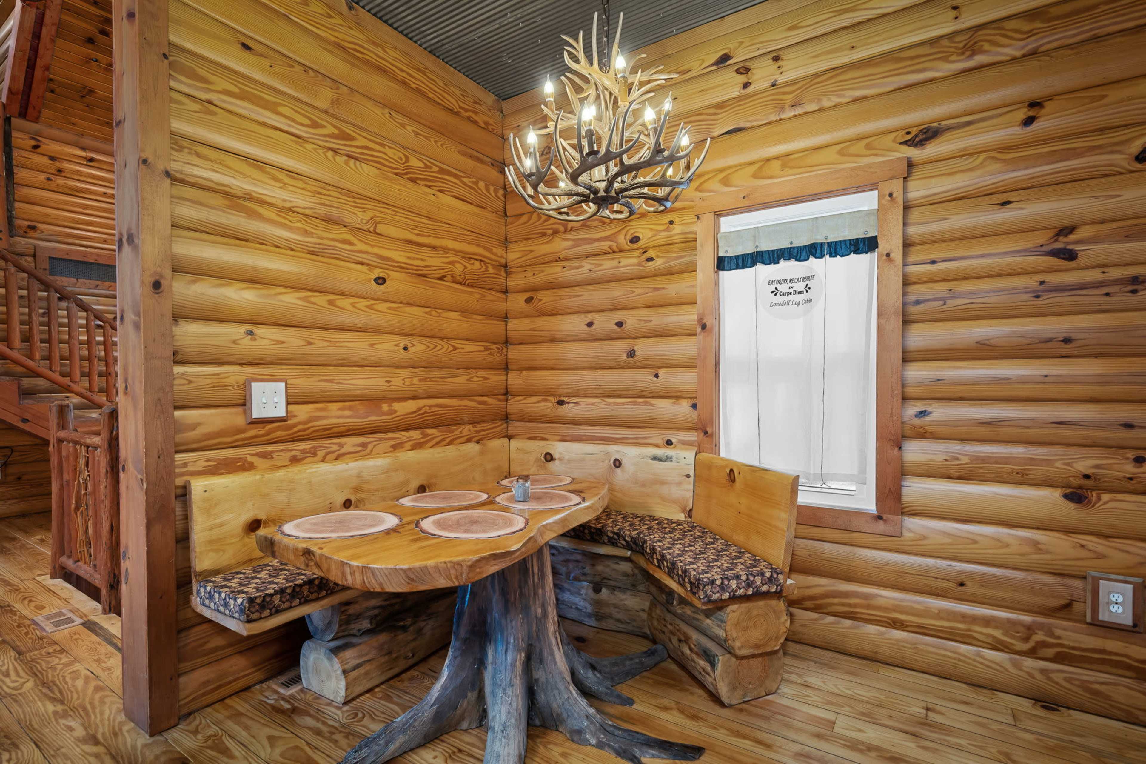 A rustic dining nook features a wooden table with a tree trunk base and a built-in bench along the wall, illuminated by an antler chandelier.