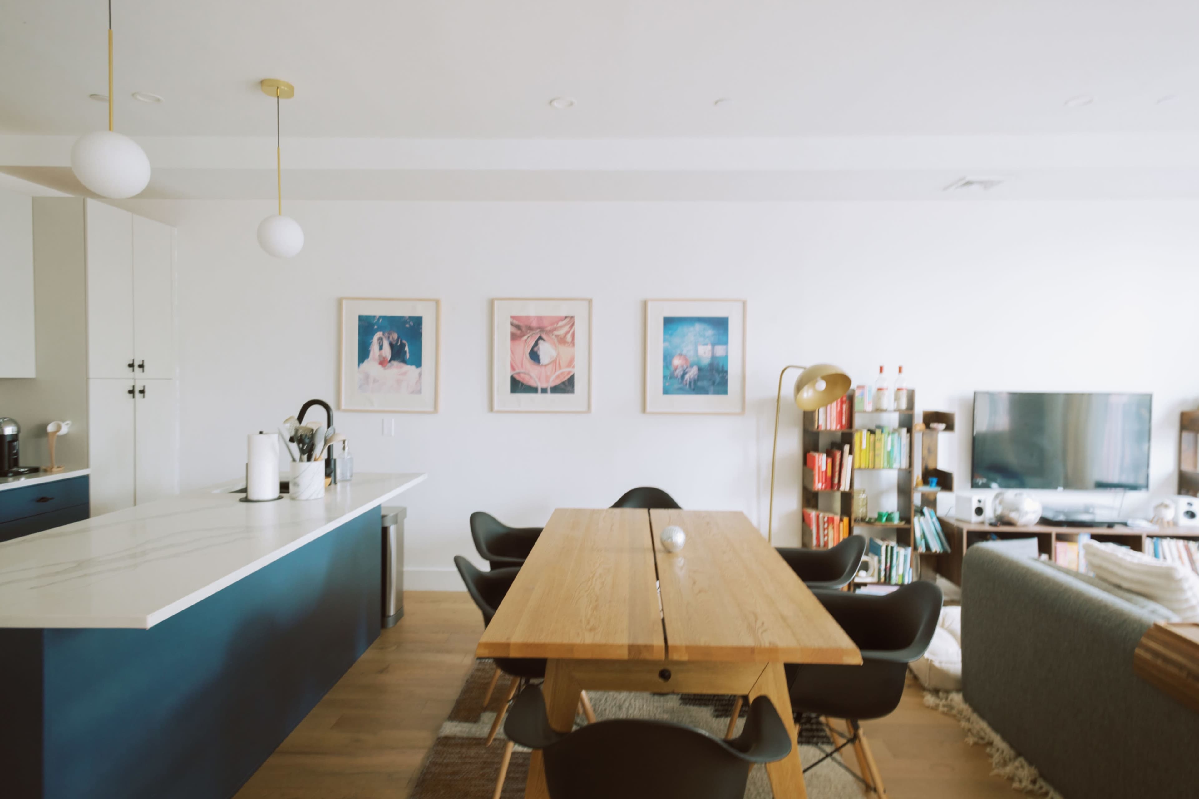 A modern kitchen and dining area features a long wooden table surrounded by black chairs, with framed artwork on the wall and a bookshelf filled with various items.