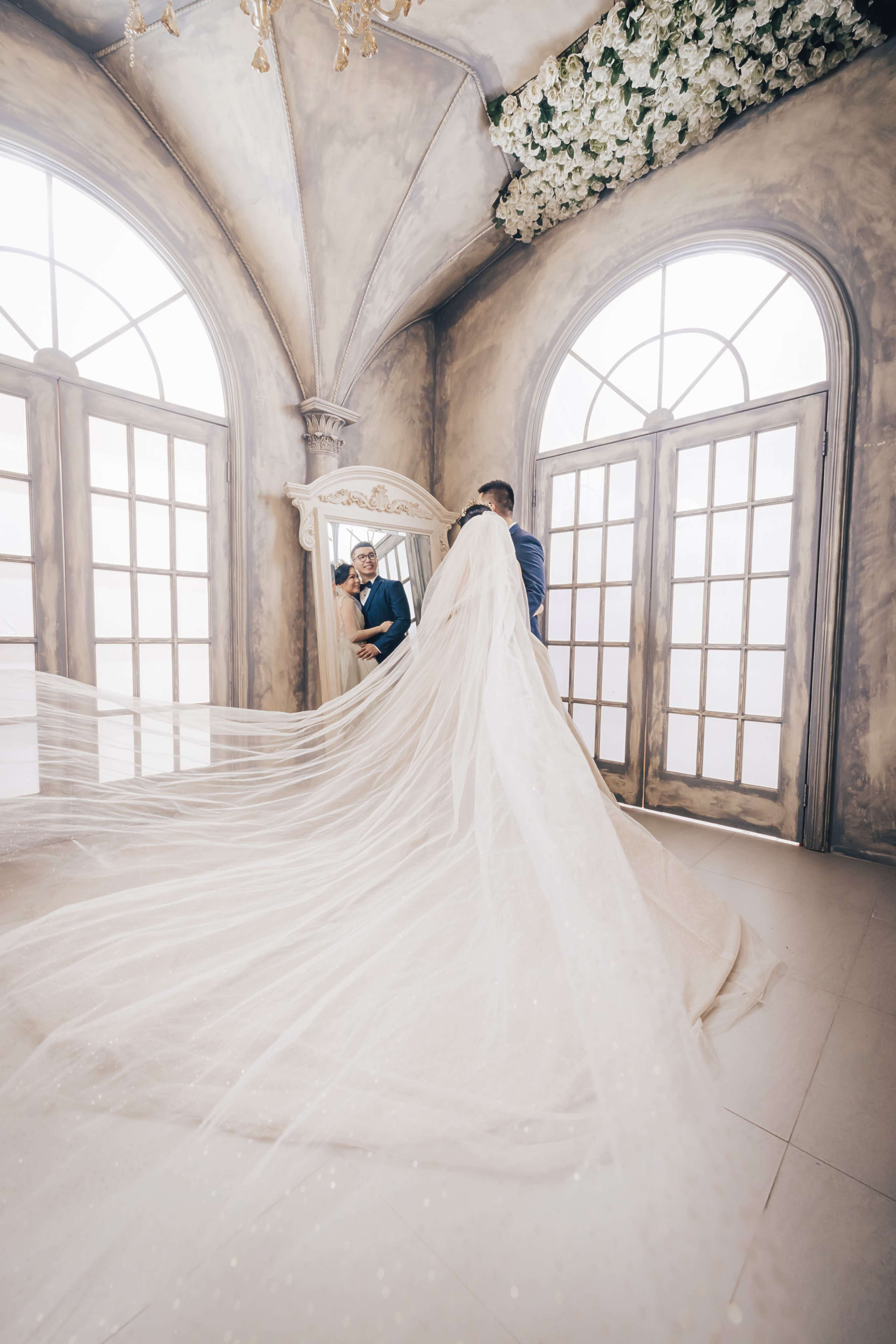 A bride in a flowing veil admires her reflection in a mirror while a groom adjusts the veil in a decorated room with large arched windows.