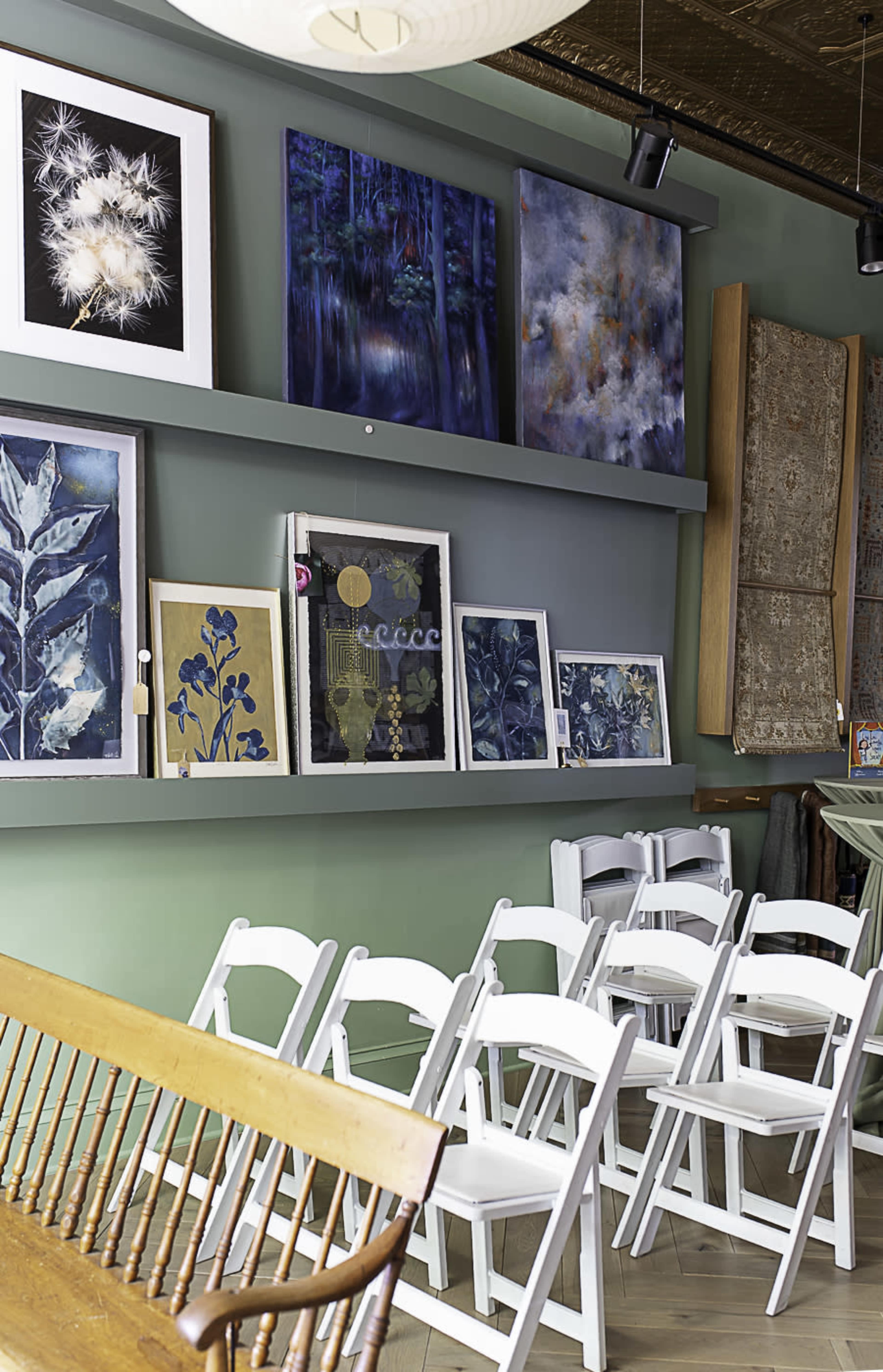 The image shows a workspace or gallery with a green wall displaying several pieces of artwork, accompanied by a row of white folding chairs positioned in front.