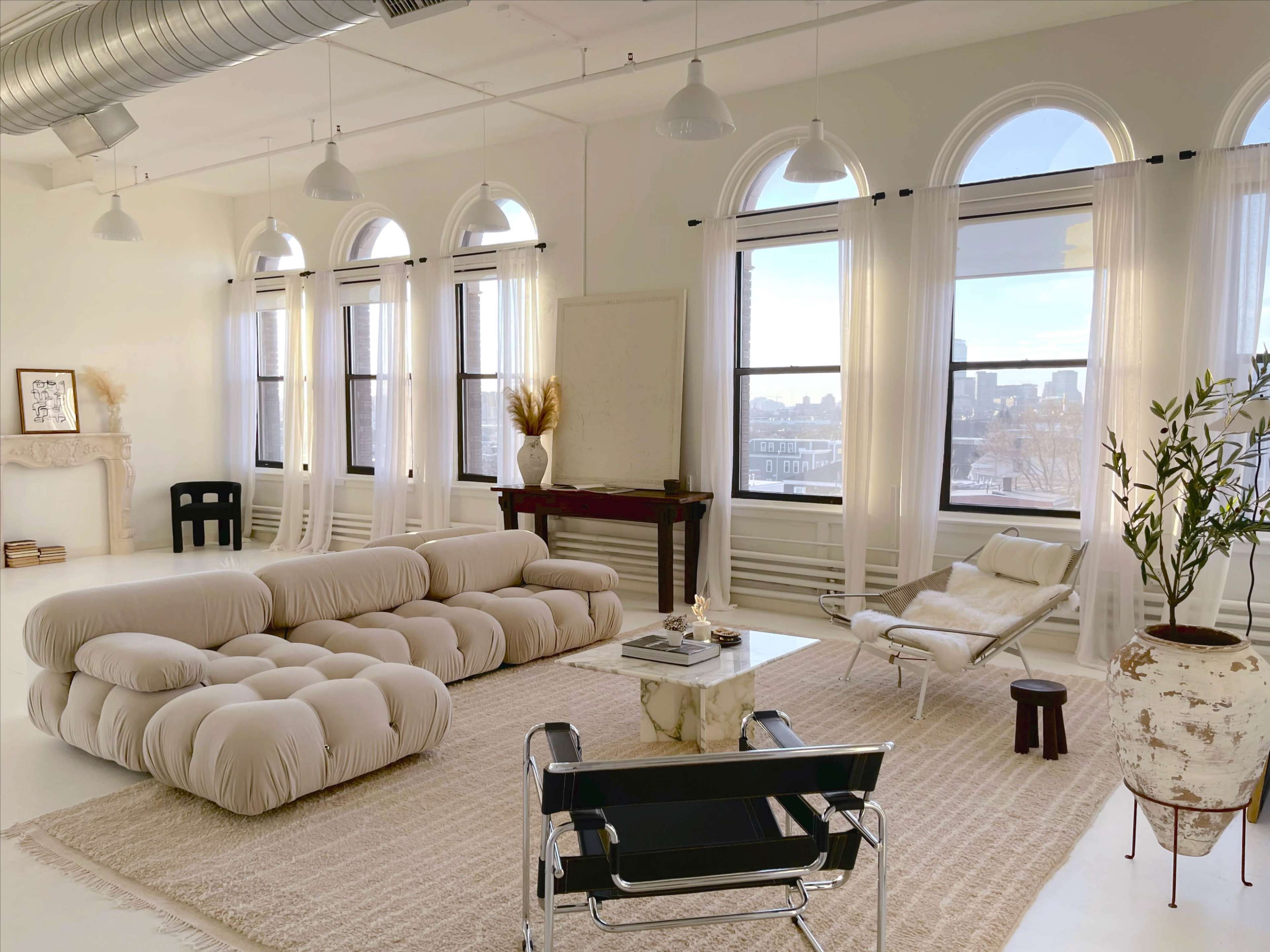 A spacious, minimalist living room featuring a plush beige sofa, large windows with sheer curtains, and a simple coffee table surrounded by modern chairs.