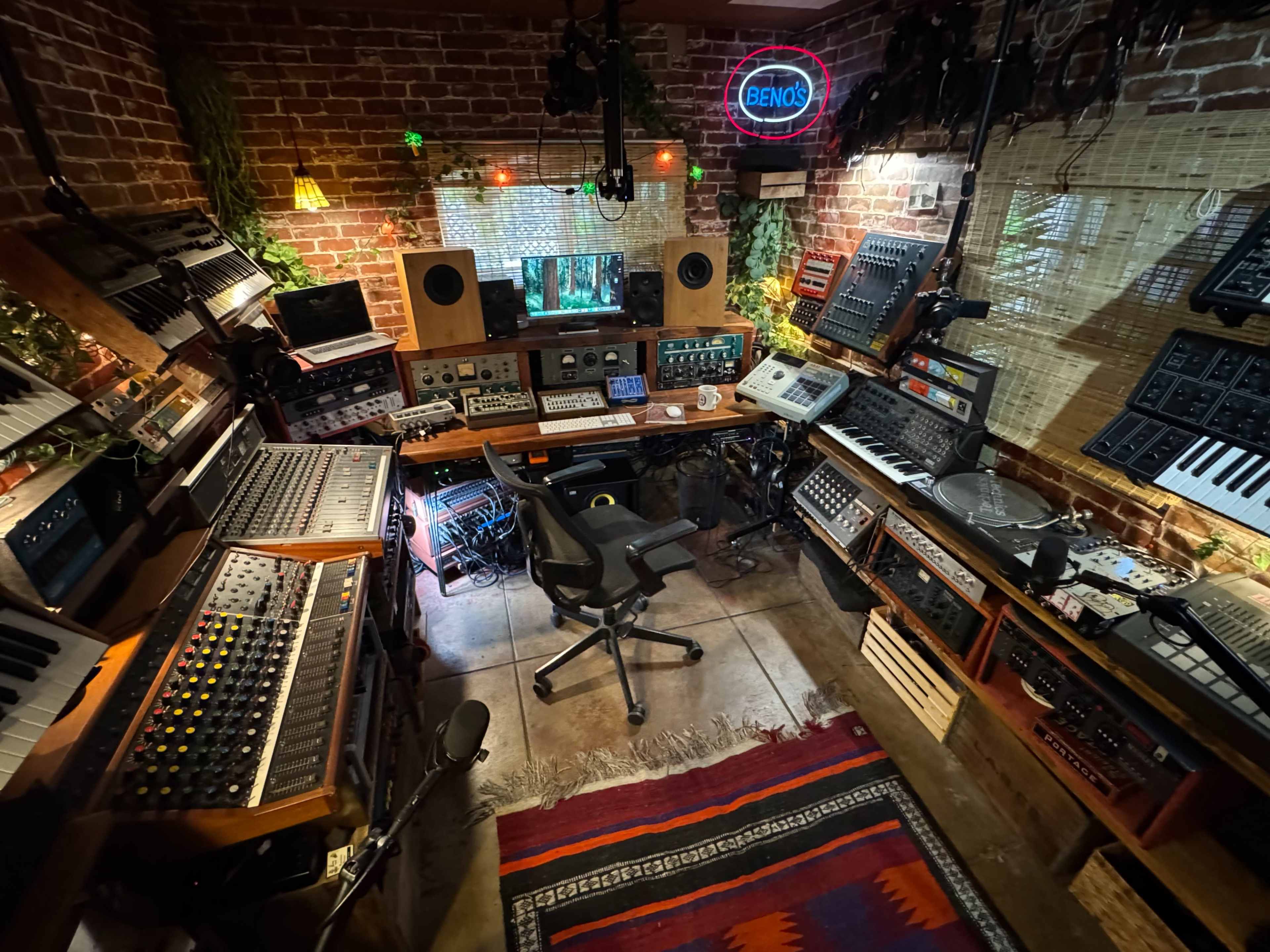 The image shows a sound production studio filled with various audio equipment, synthesizers, and a desktop setup under dim lighting.