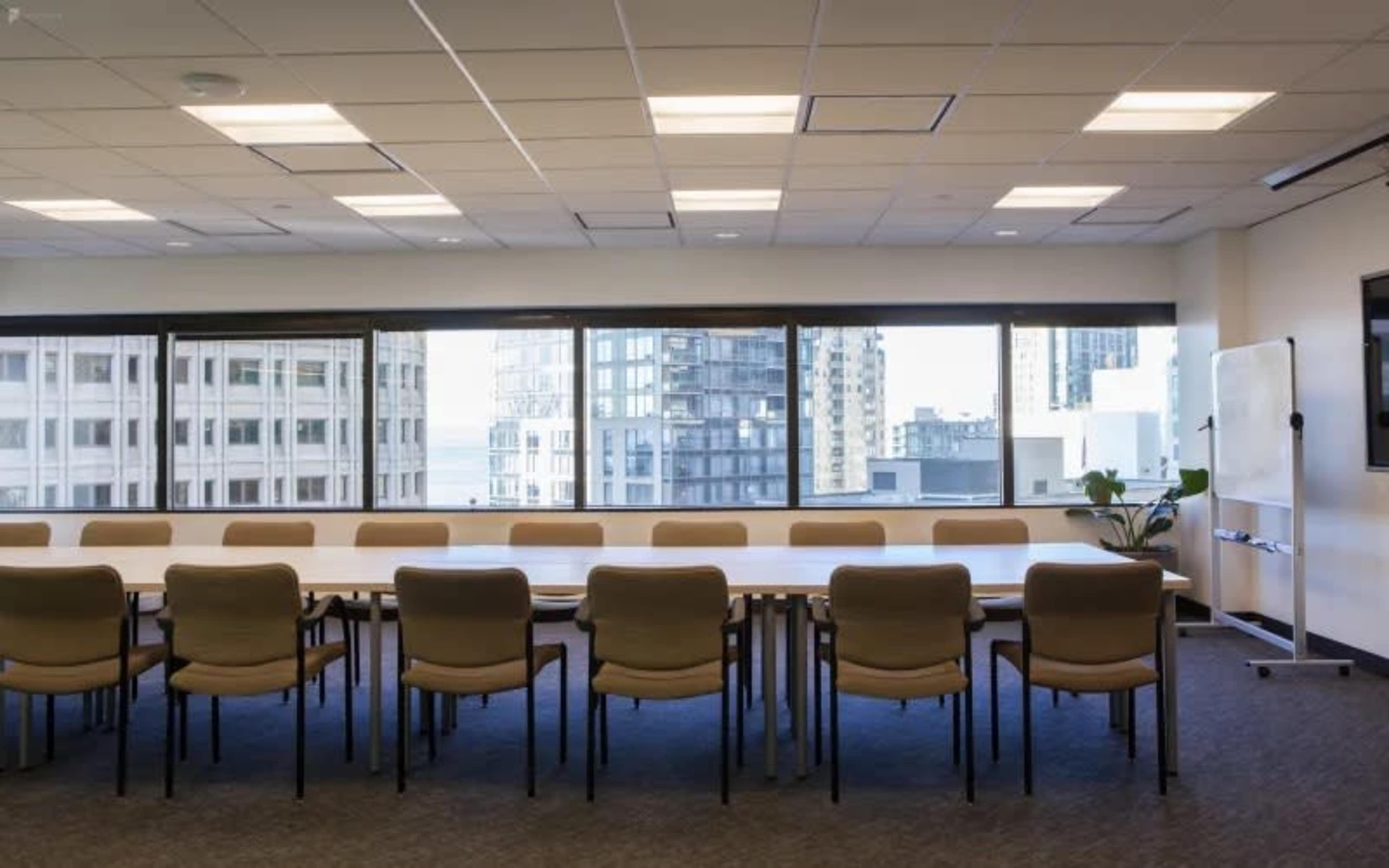A large conference room features a long table surrounded by chairs, with large windows offering a view of city buildings.