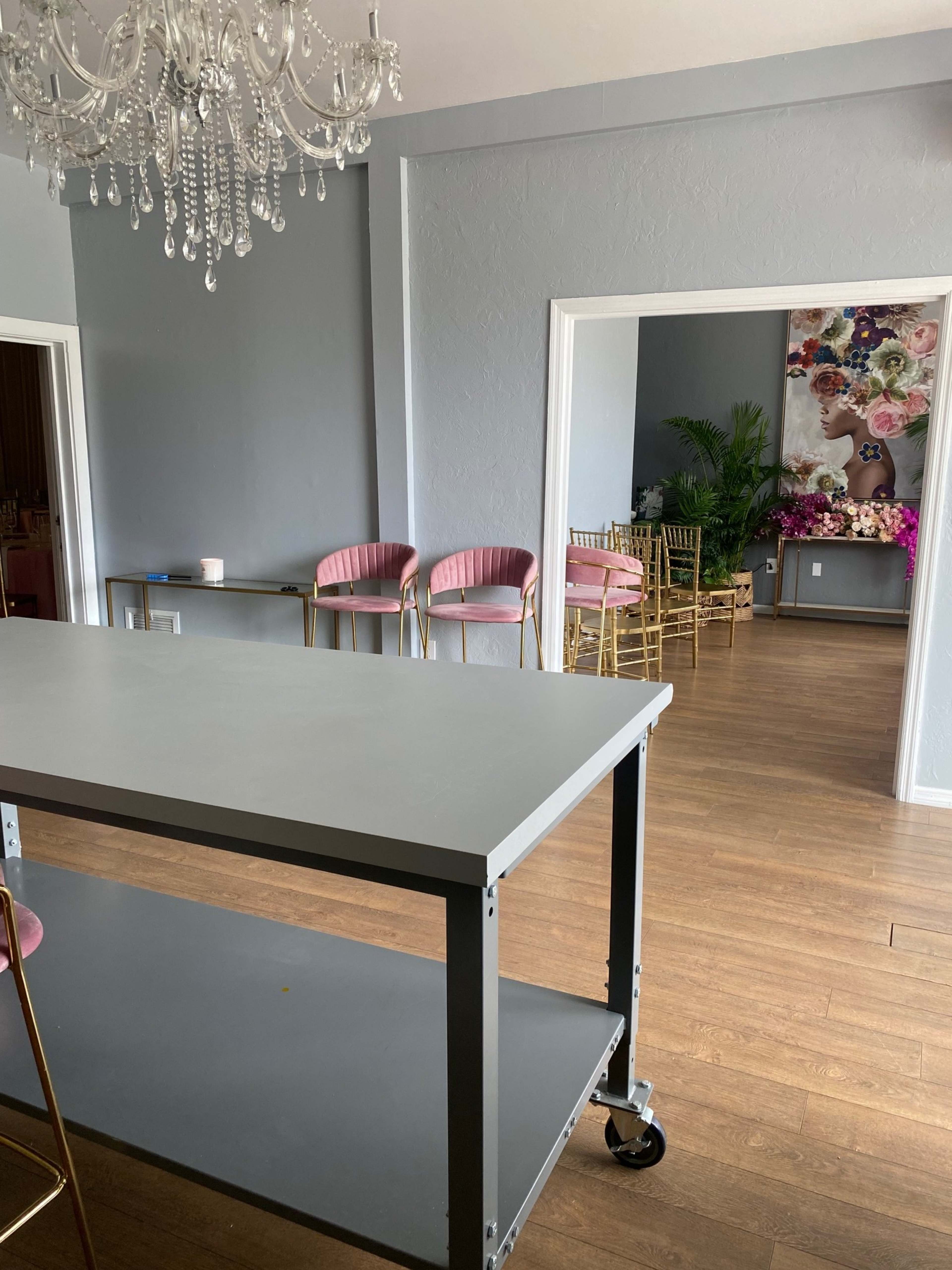 The image shows a spacious room with a modern table on wheels in the foreground and pink chairs in the background, illuminated by a chandelier and featuring decorative floral artwork on the wall.
