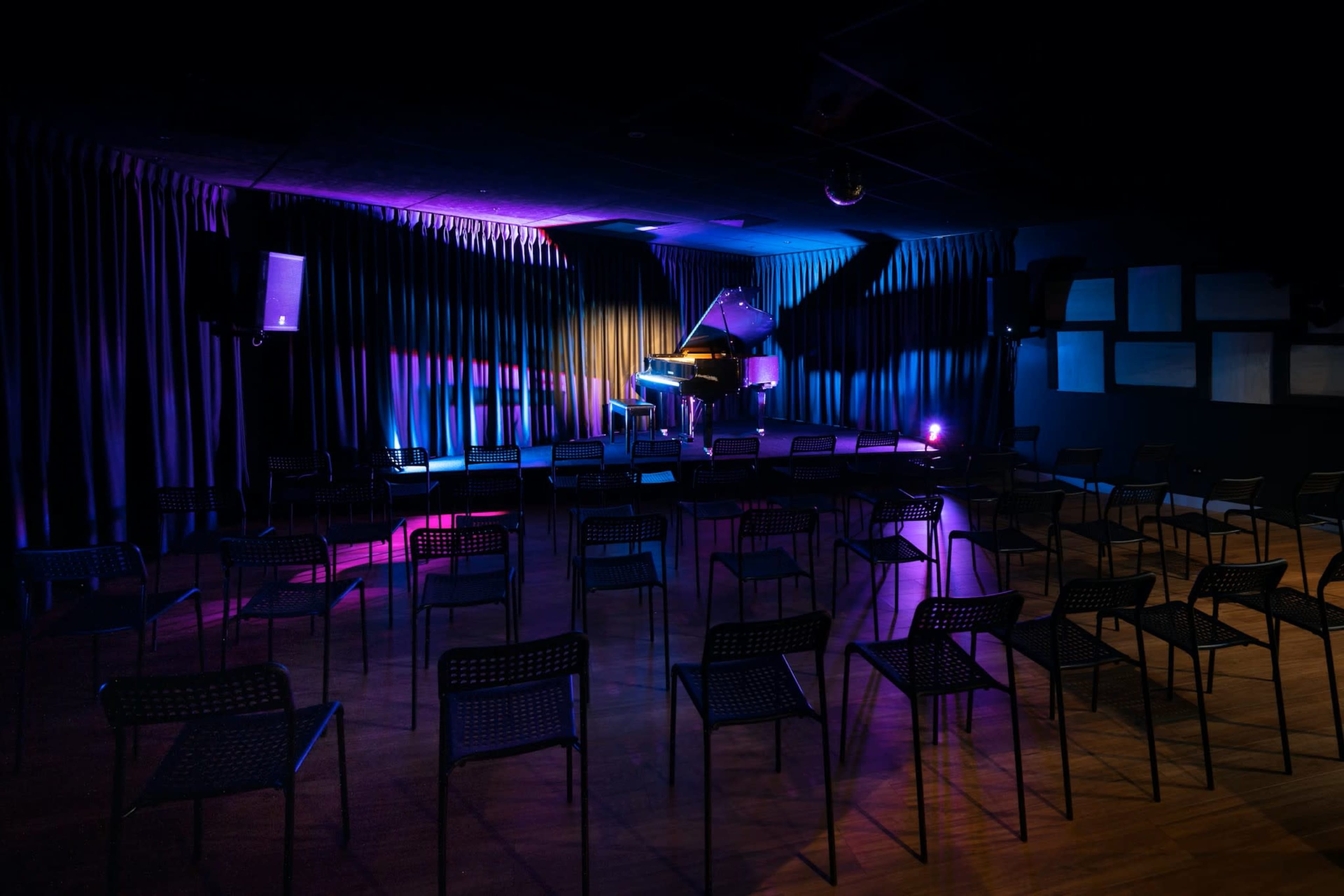 A dimly lit performance venue features a grand piano at the center, surrounded by empty black chairs arranged in rows.