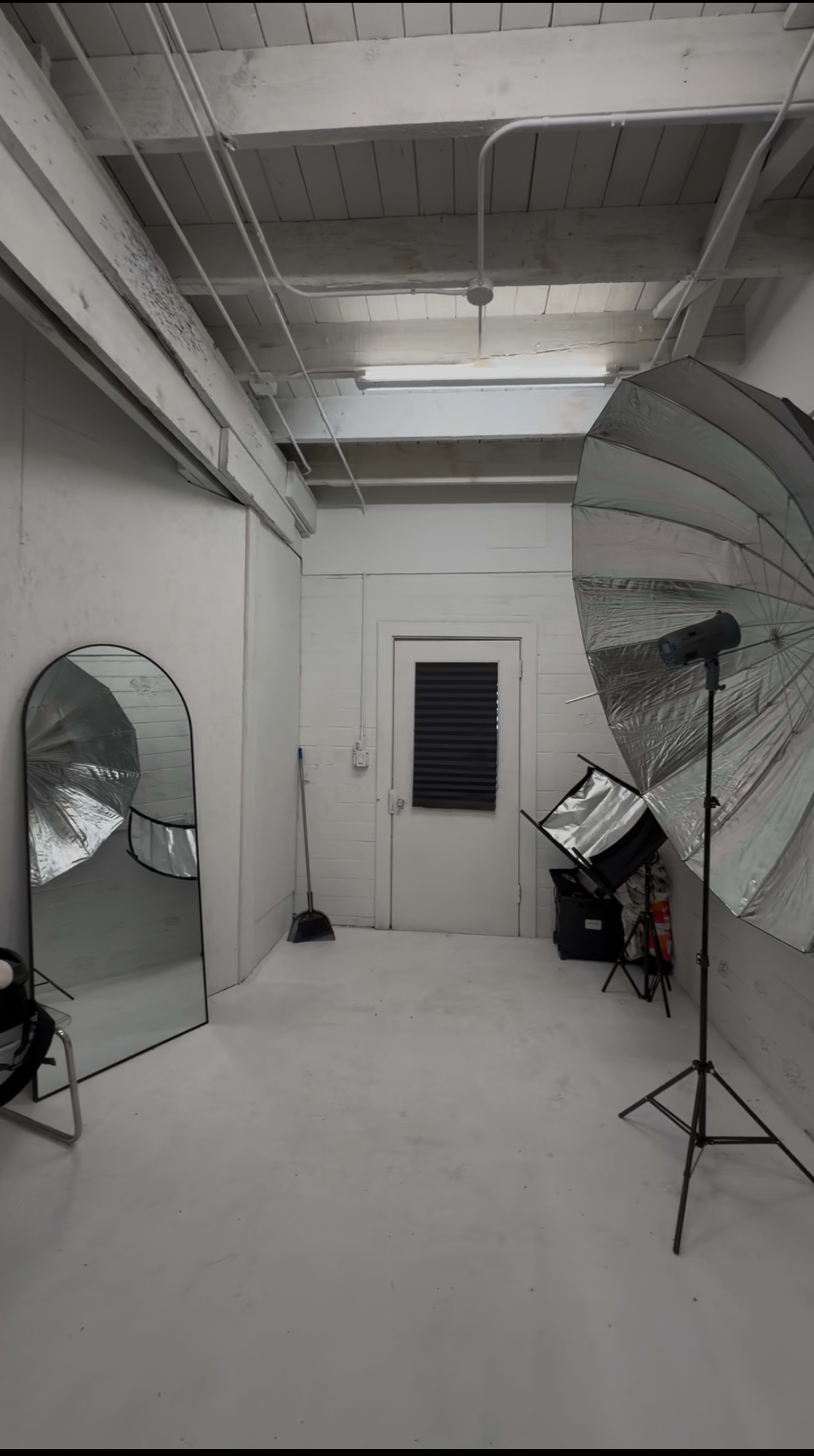 The image shows a spacious, minimalist photo studio with a large mirror, a photography umbrella reflector on a stand, and various equipment placed around the room.