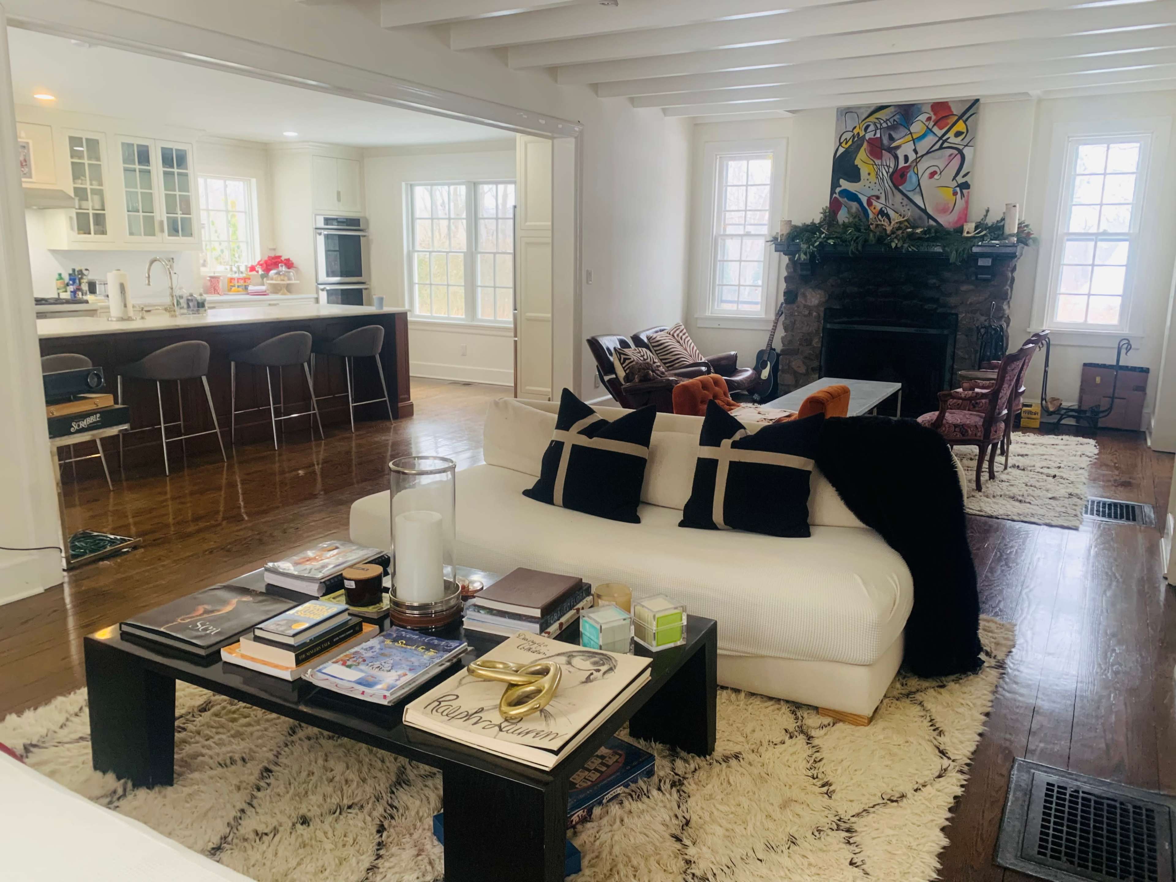 A living room features a white sofa with black pillows, a coffee table stacked with books, and a stone fireplace, with a view of a kitchen and dining area in the background.