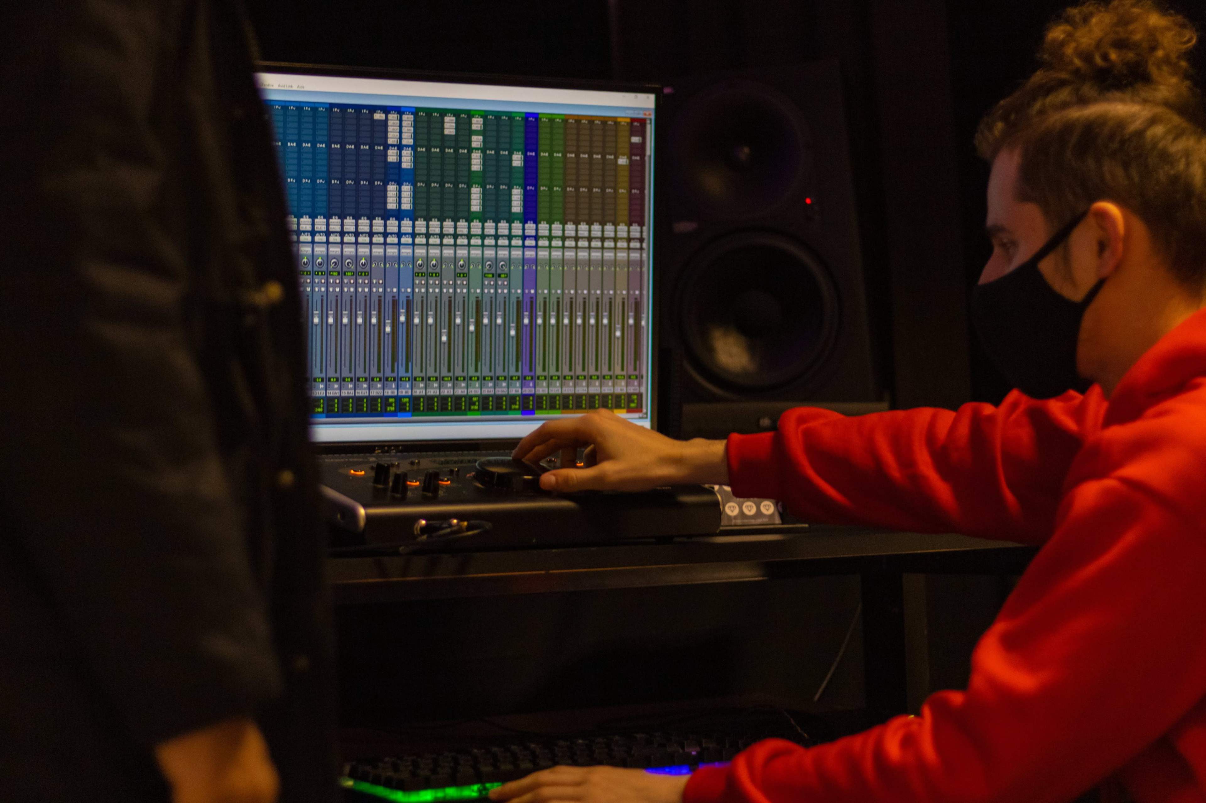 A person in a red sweatshirt works on a sound mixing software on a computer screen while another figure stands nearby.