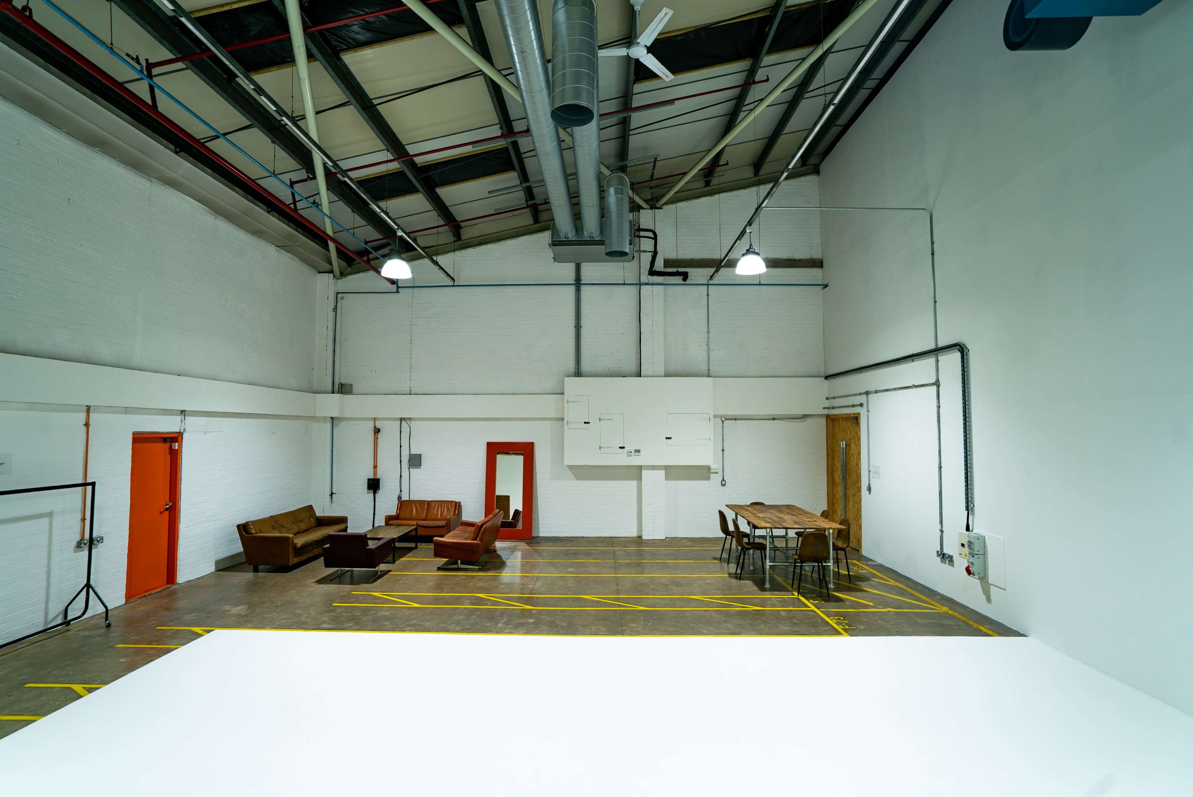 The image shows a spacious, well-lit industrial room featuring a seating area with brown couches and a long wooden table against a stark white wall.
