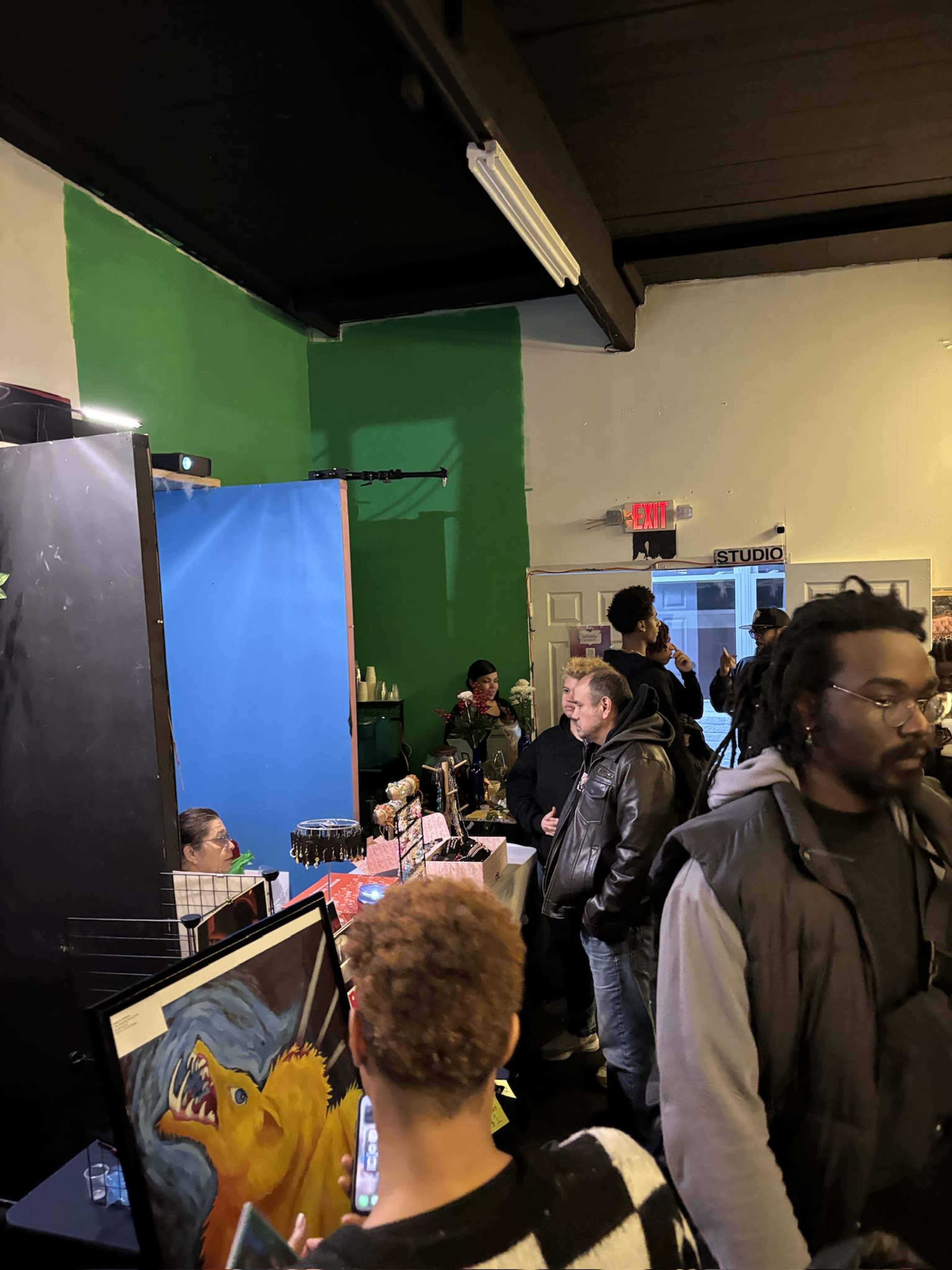 A group of people is gathered inside a room with green and blue walls, where art displays and a vendor table are visible.