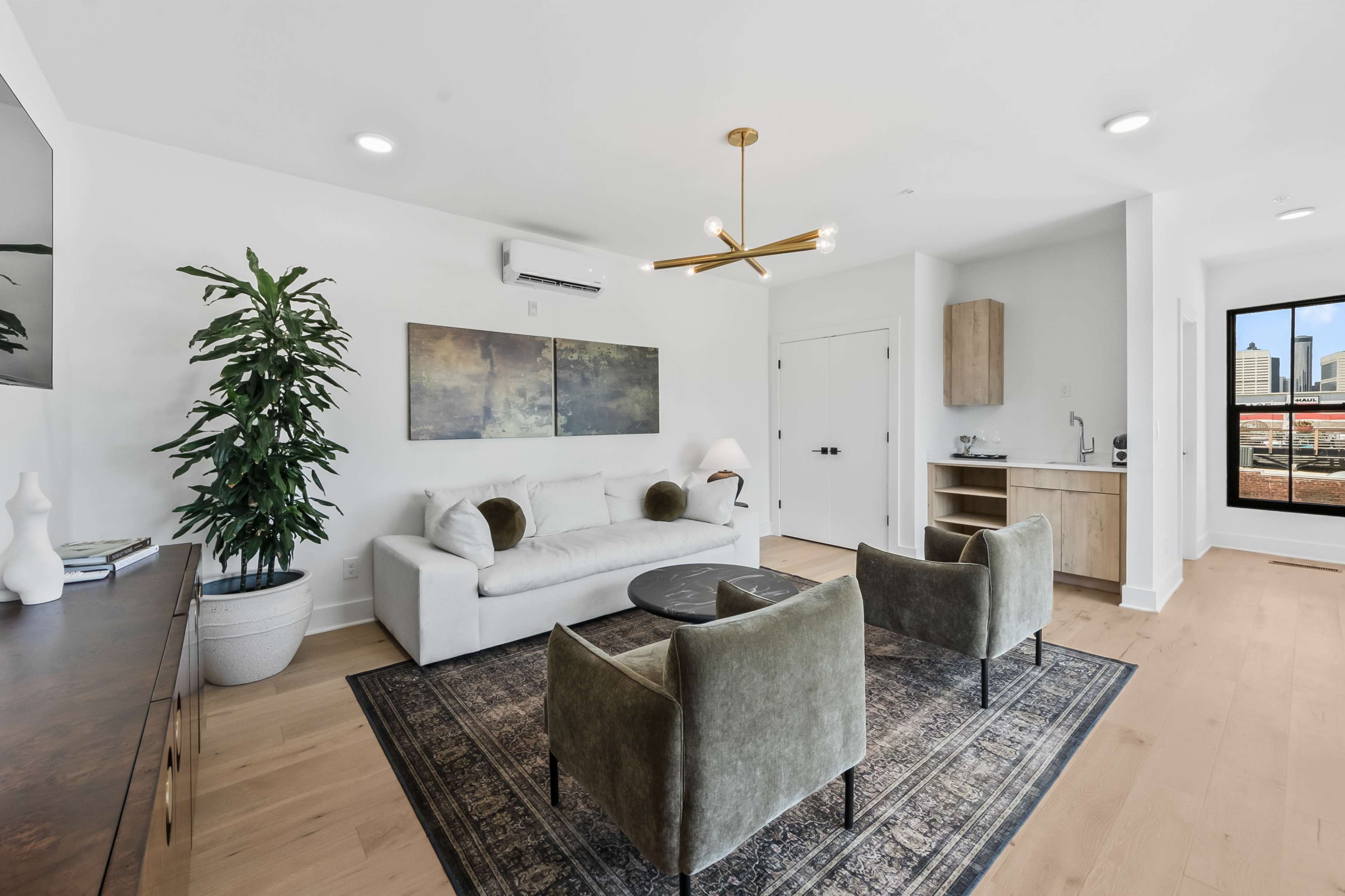 The image shows a modern living room featuring a white sofa, two green armchairs, a glass coffee table, and a kitchenette in the background, with a large window revealing a city view.