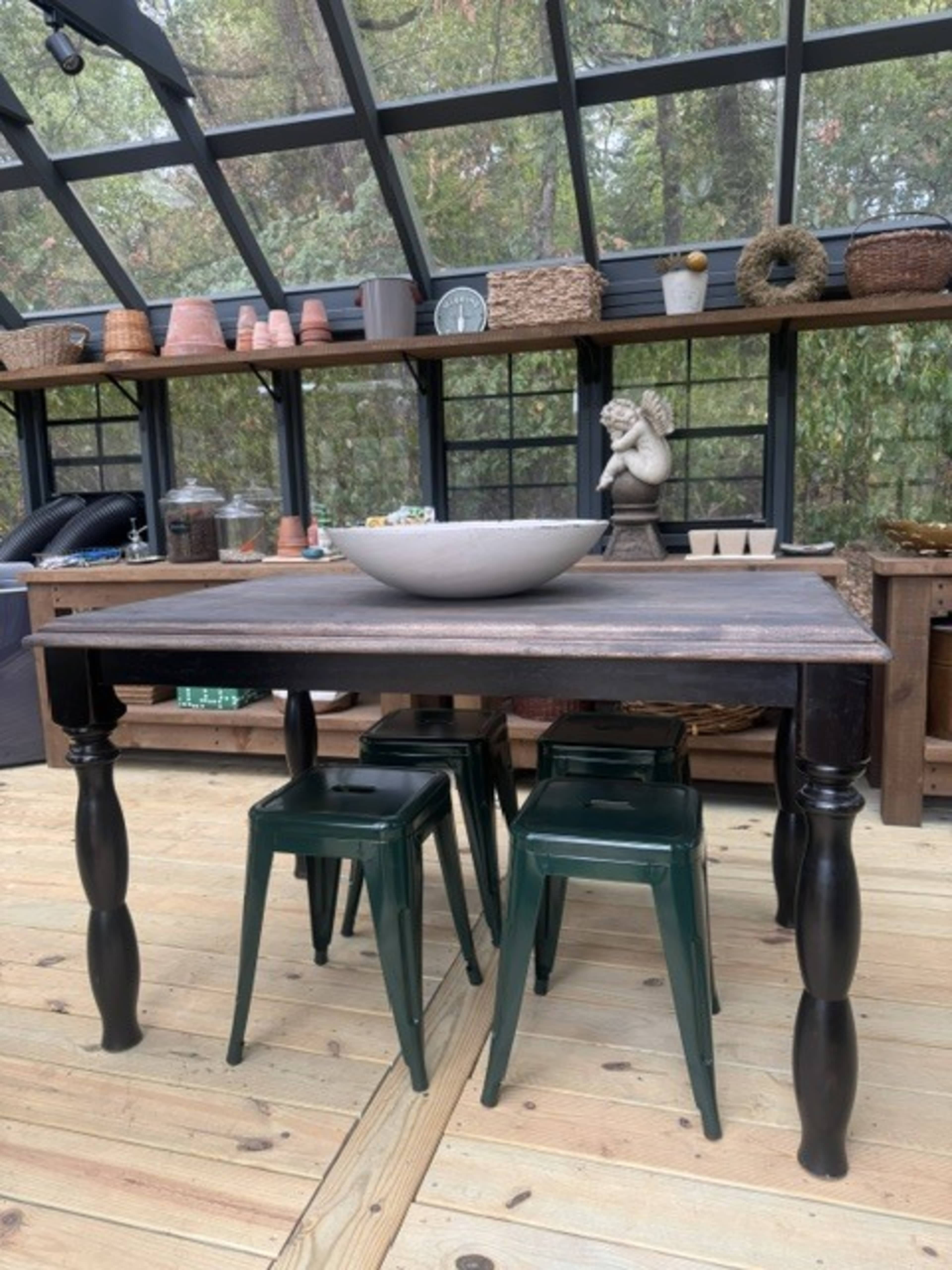 A wooden table with turned legs is set in a sunroom, surrounded by green metal stools and various decor items on the shelves.