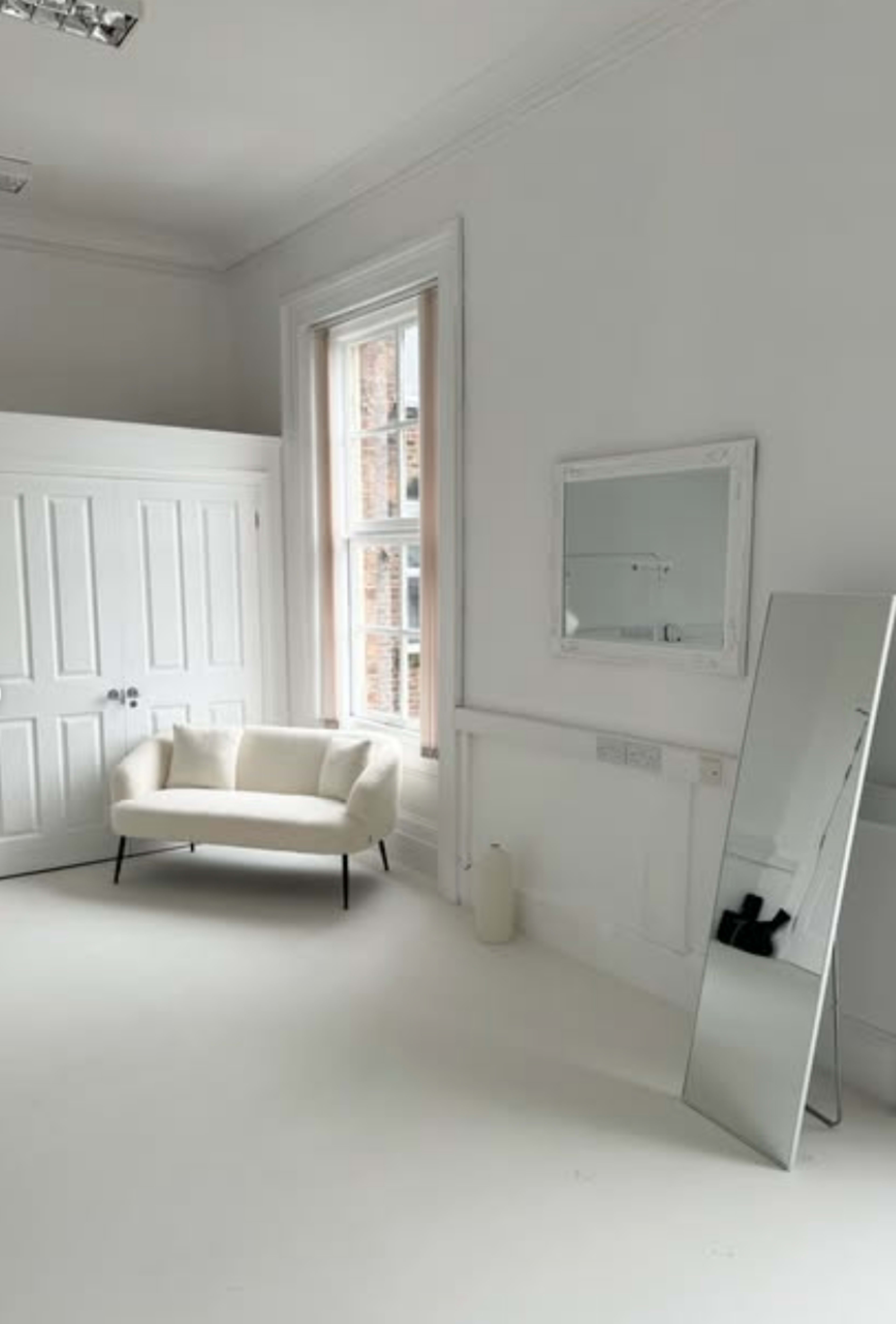 The image shows a minimalist room with white walls, a large mirror, a light-colored sofa, and a window alongside a closet with double doors.