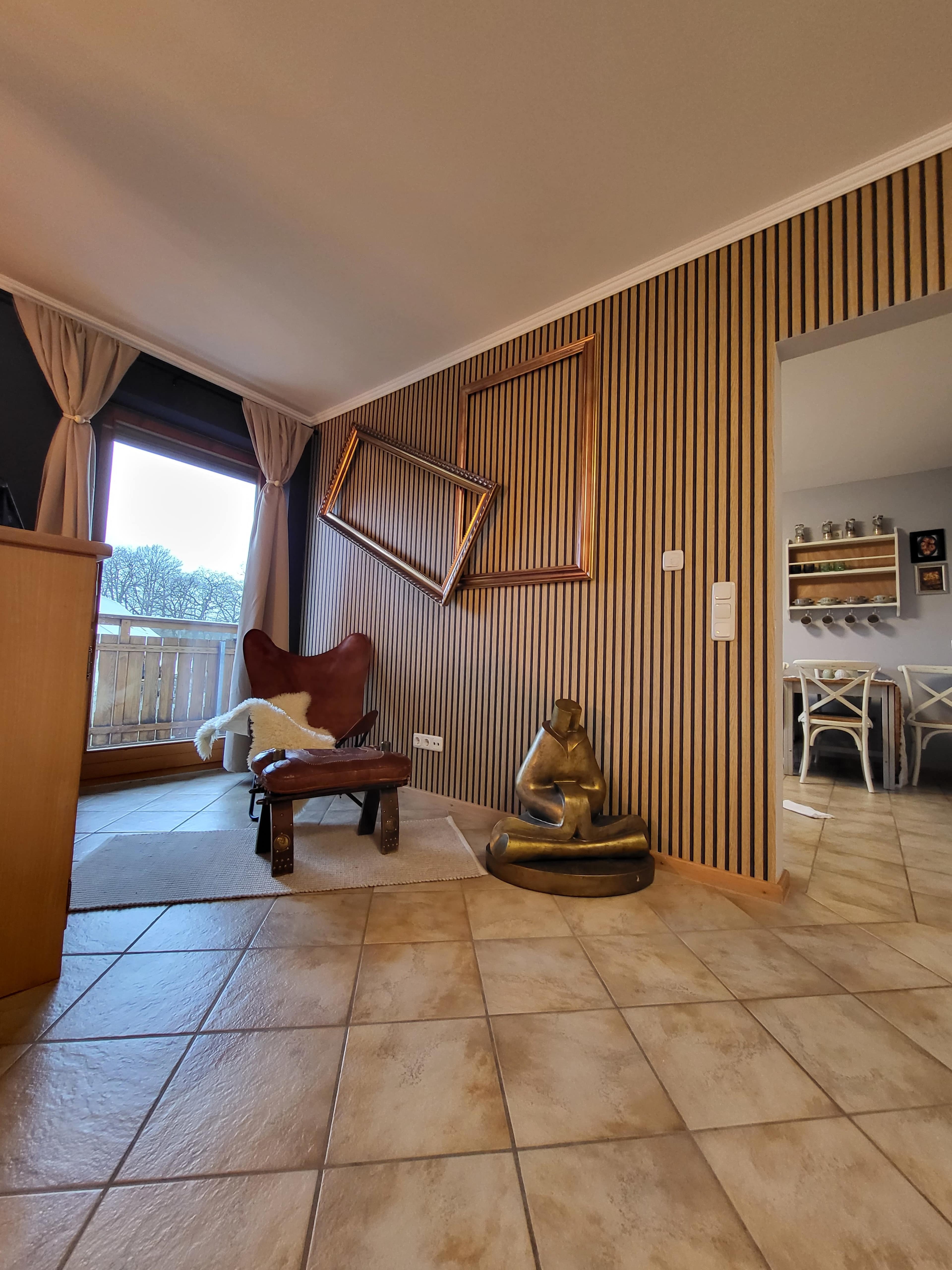 A corner of a room with a wooden floor, a leather chair, a statue of a seated figure, and a tilted picture frame mounted on a striped wall.