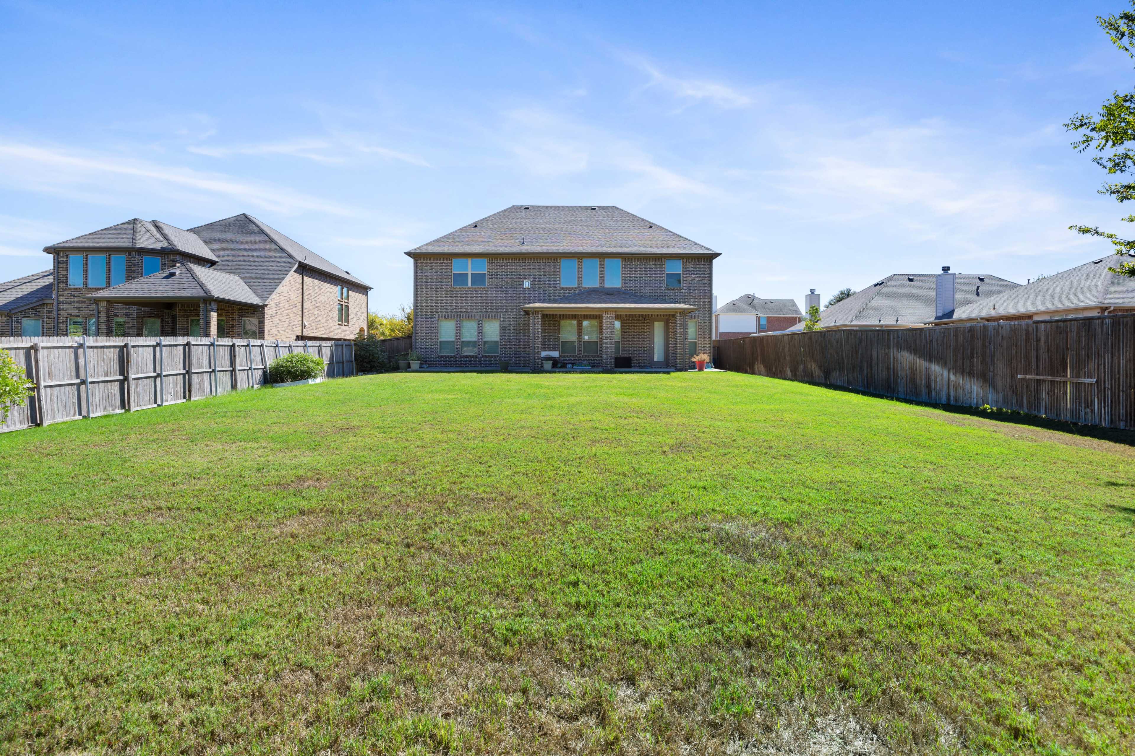 The image shows a spacious backyard with freshly mowed grass, bordered by wooden fences and two two-story houses in the background.