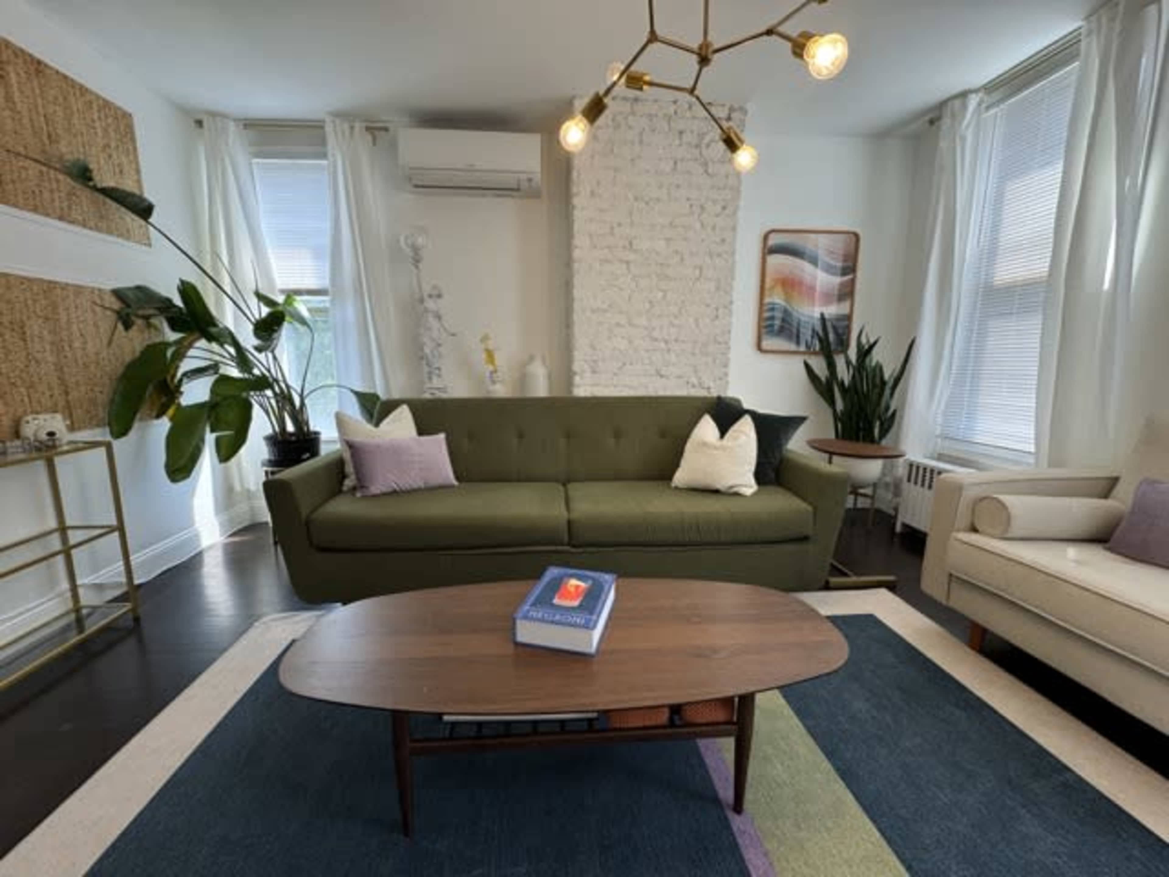 The image shows a cozy living room with a green sofa, a wooden coffee table, and a mix of plants and decorative elements against a white brick wall.