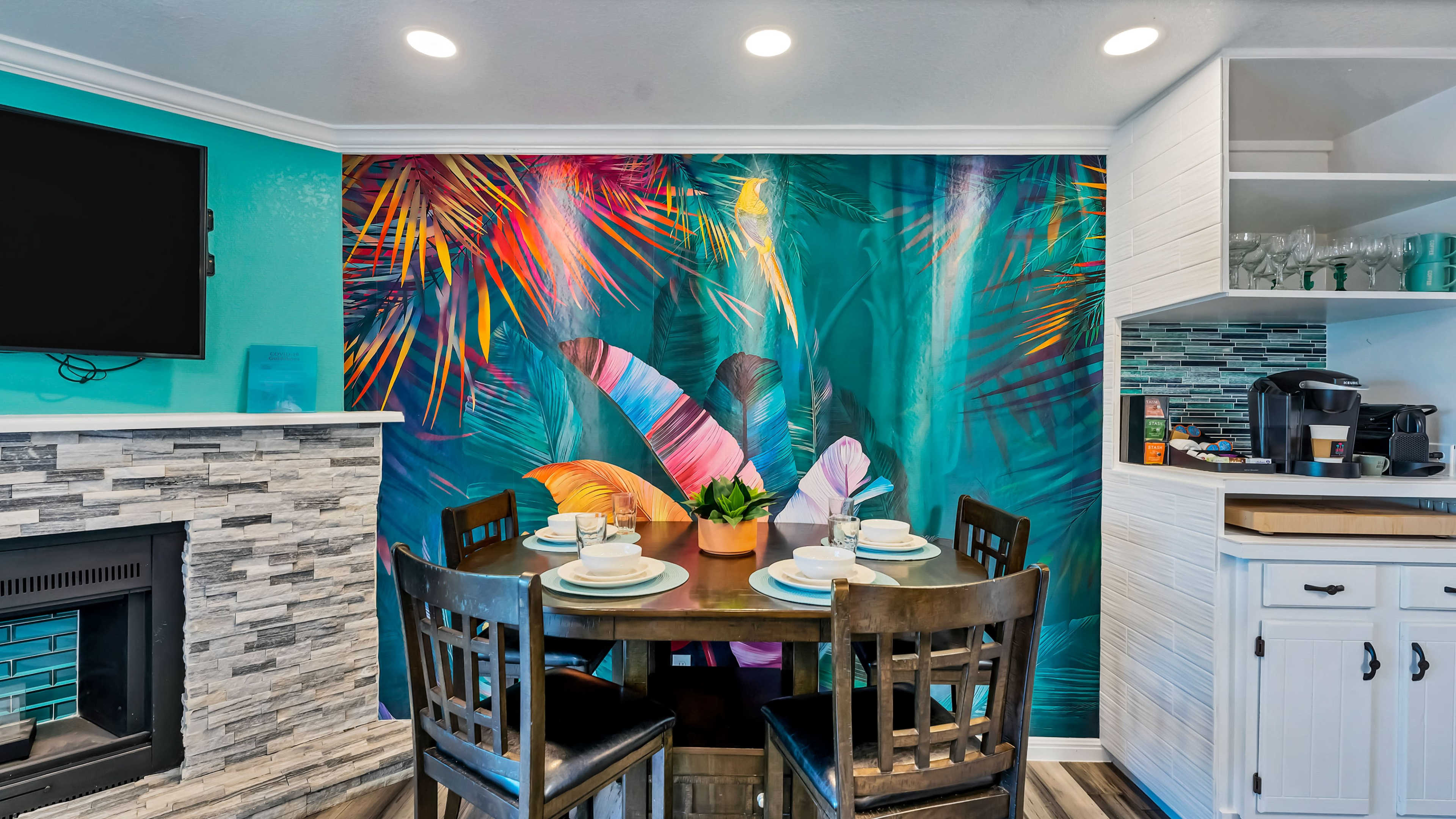 A dining area features a round table set for four, with a vibrant tropical mural on the wall and a modern kitchenette nearby.