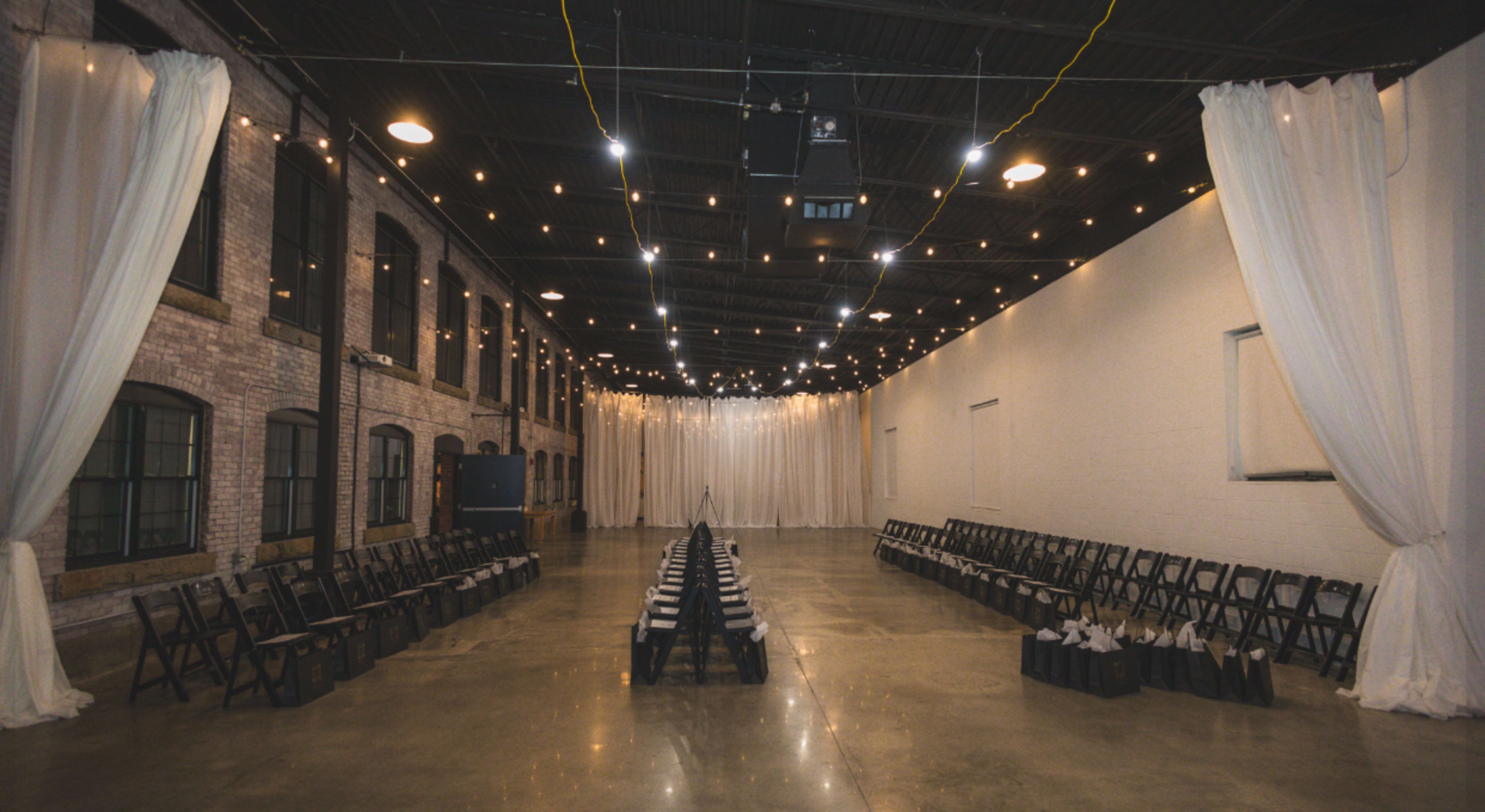 The image shows an indoor venue with rows of black chairs facing a central aisle, illuminated by string lights and adorned with white drapes along the walls.