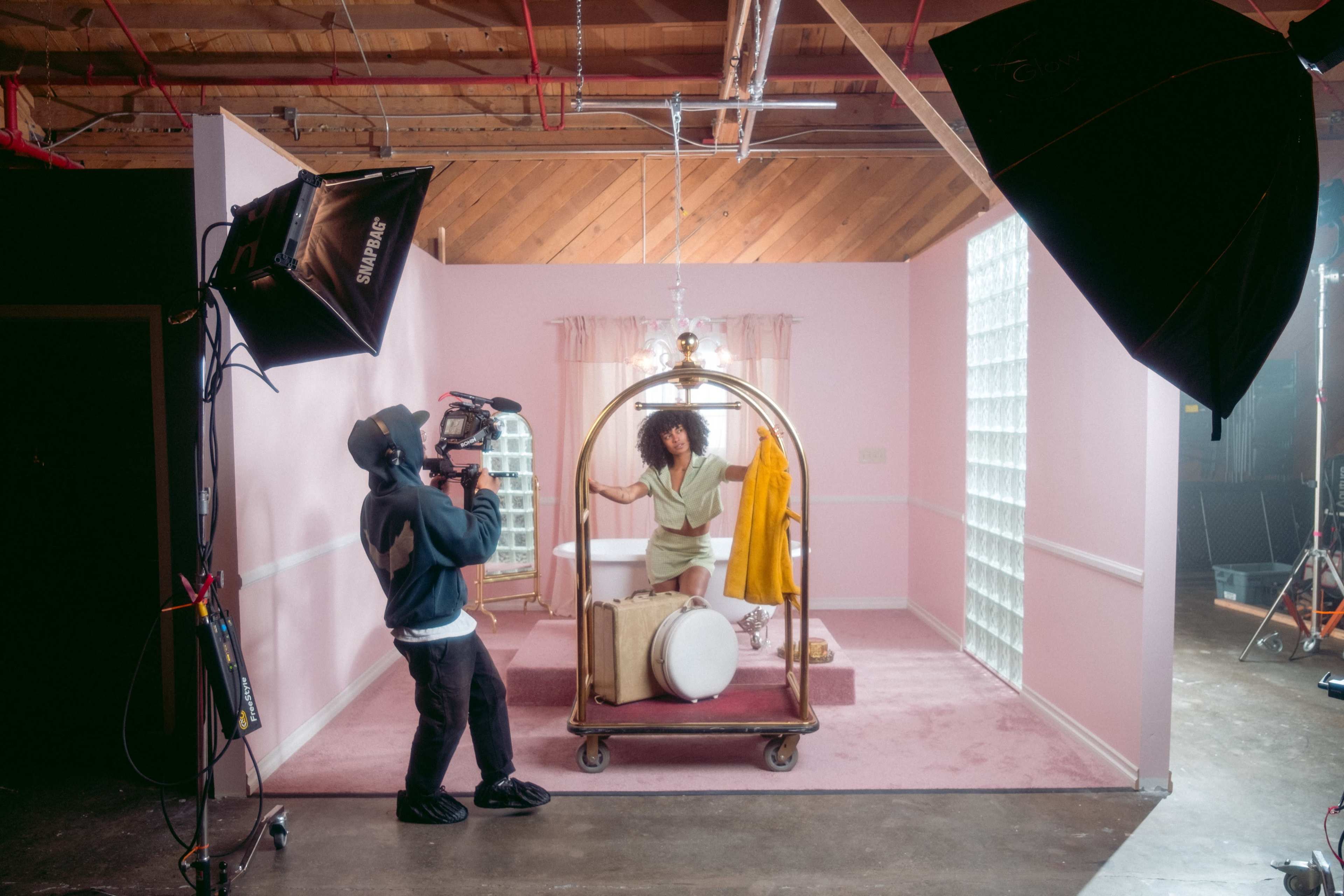 A woman poses inside a vintage bathtub on a pink set while a cameraman captures the scene with a professional camera.
