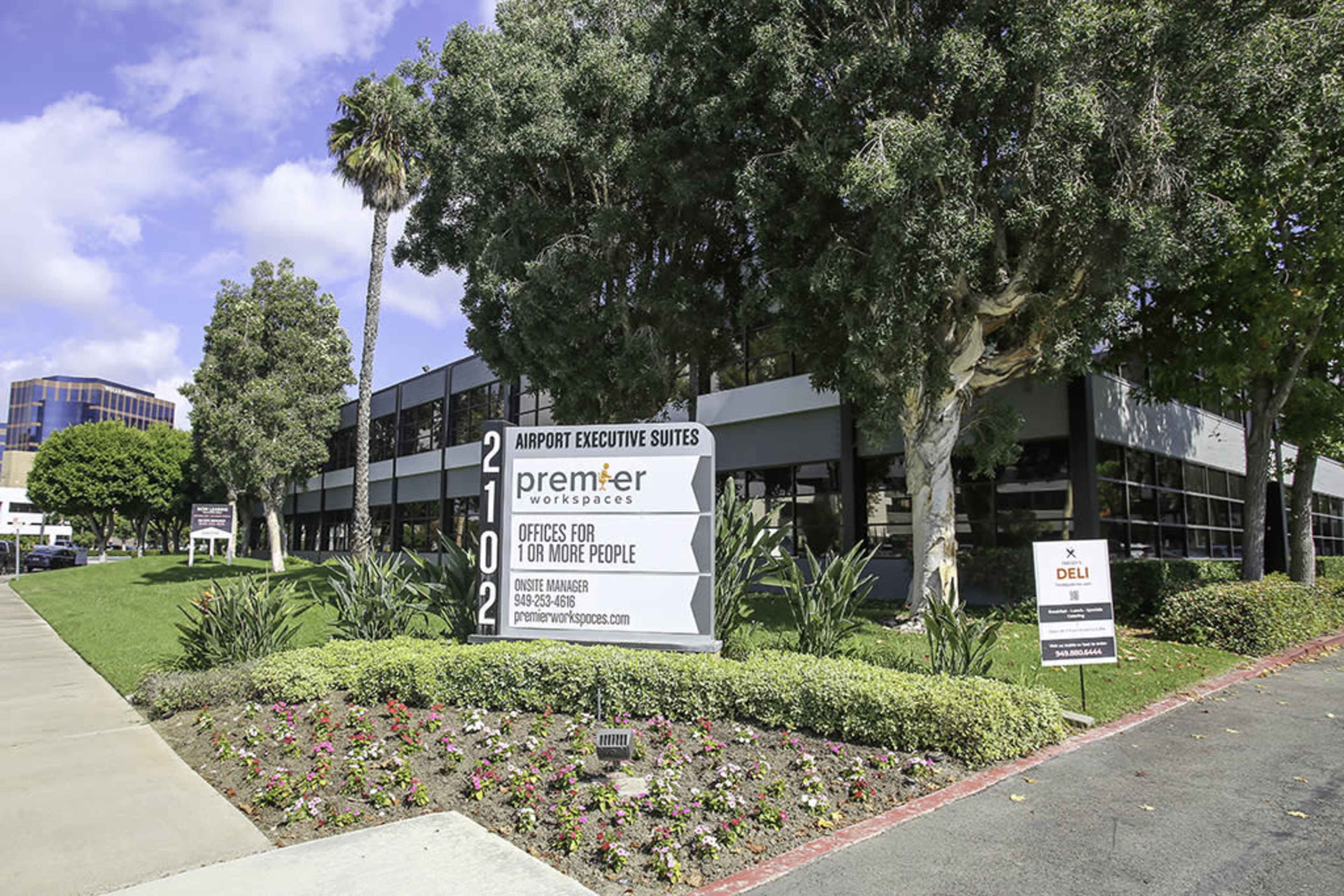 The image shows a modern building with a sign for "Airport Executive Suites" and landscaping featuring flowers and palm trees.