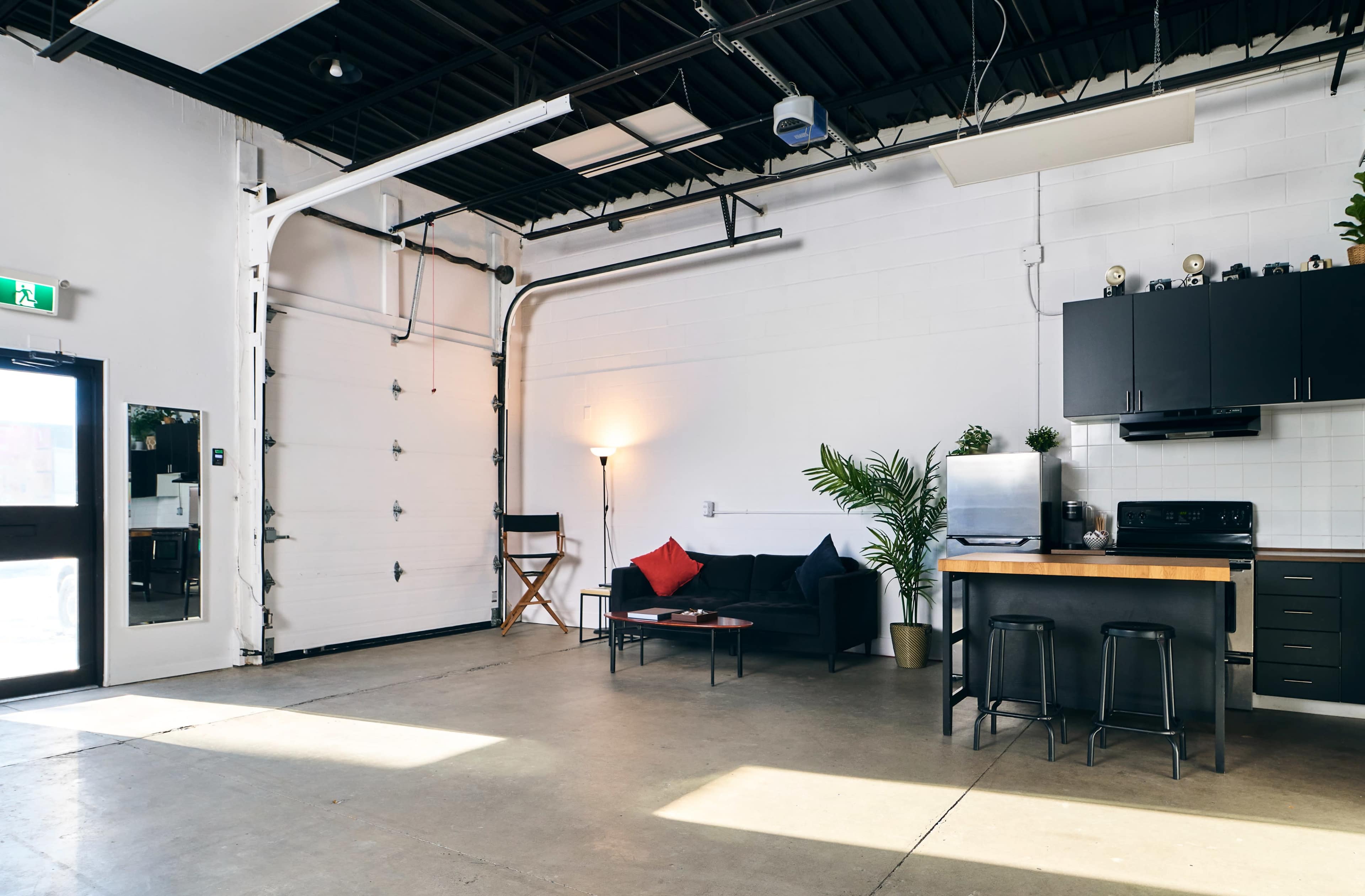 The image shows a spacious room with a minimalist design, featuring a couch, a kitchen island, a lamp, and a plant, illuminated by natural light from the large windows.