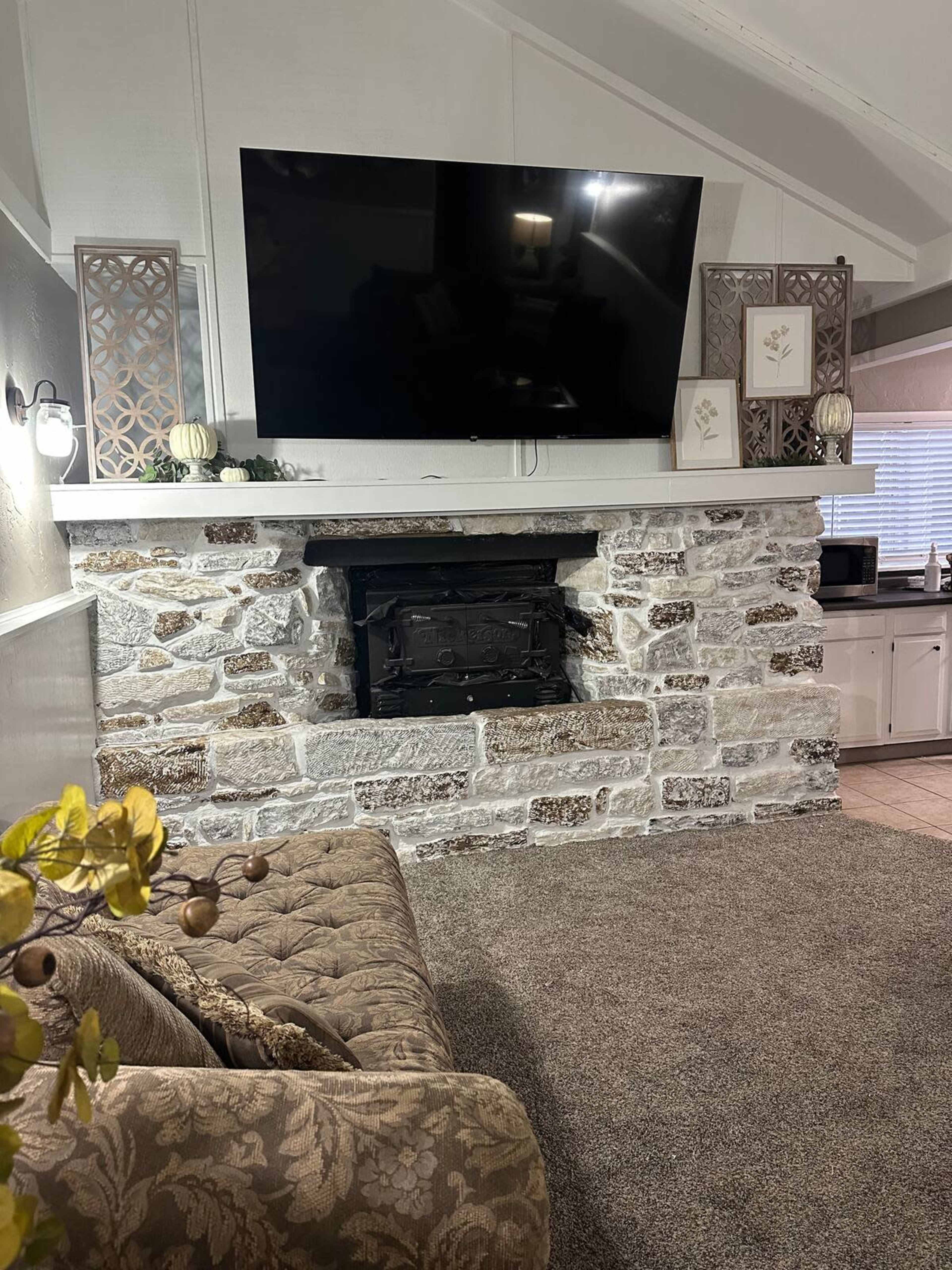 The image shows a living room with a stone fireplace beneath a large flat-screen TV, accented by decorative wall features and a plush sofa in the foreground.