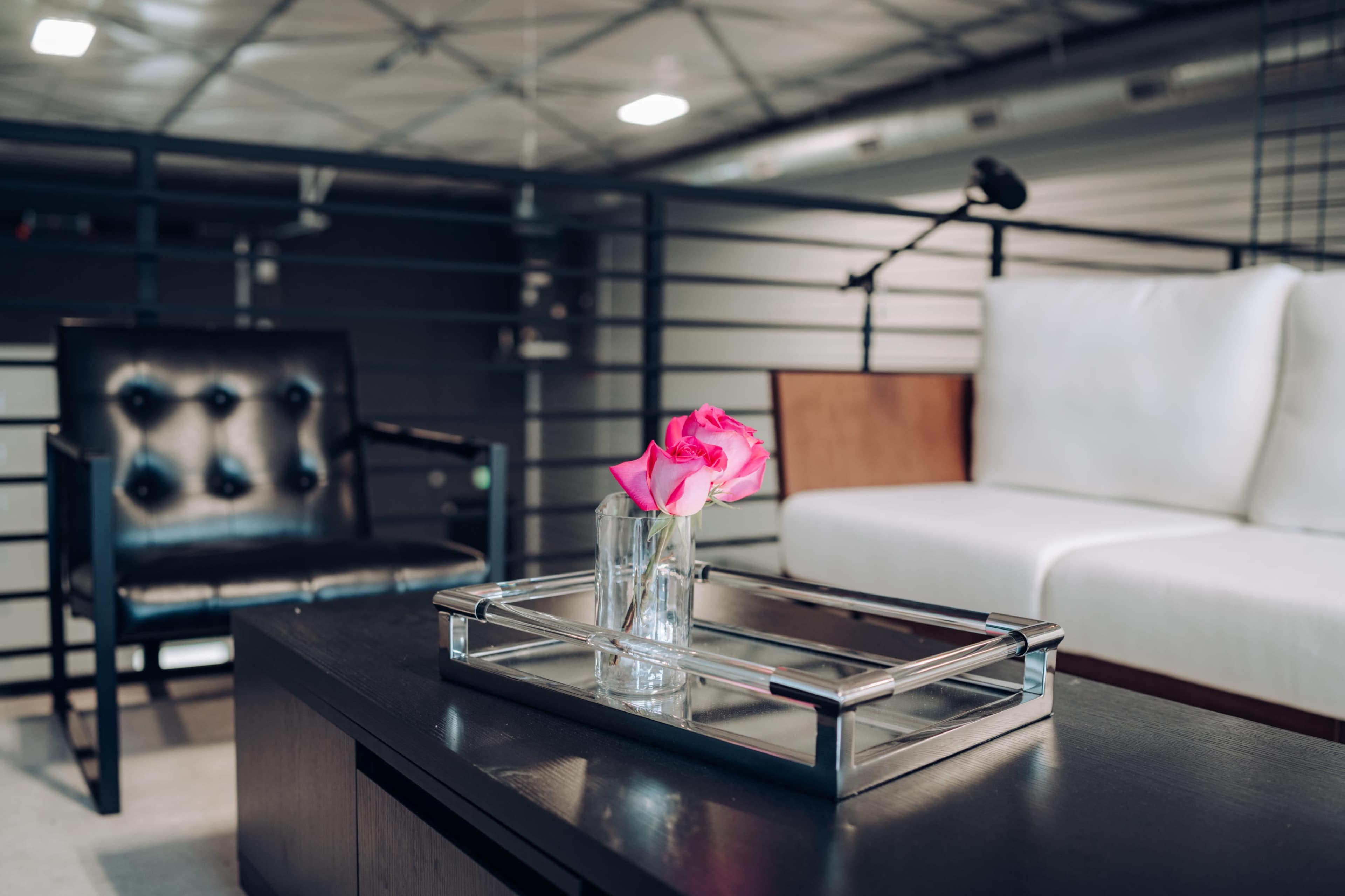 A glass vase with a pink flower sits on a mirrored tray on a black coffee table in a modern lounge area featuring a black chair and a white sofa.