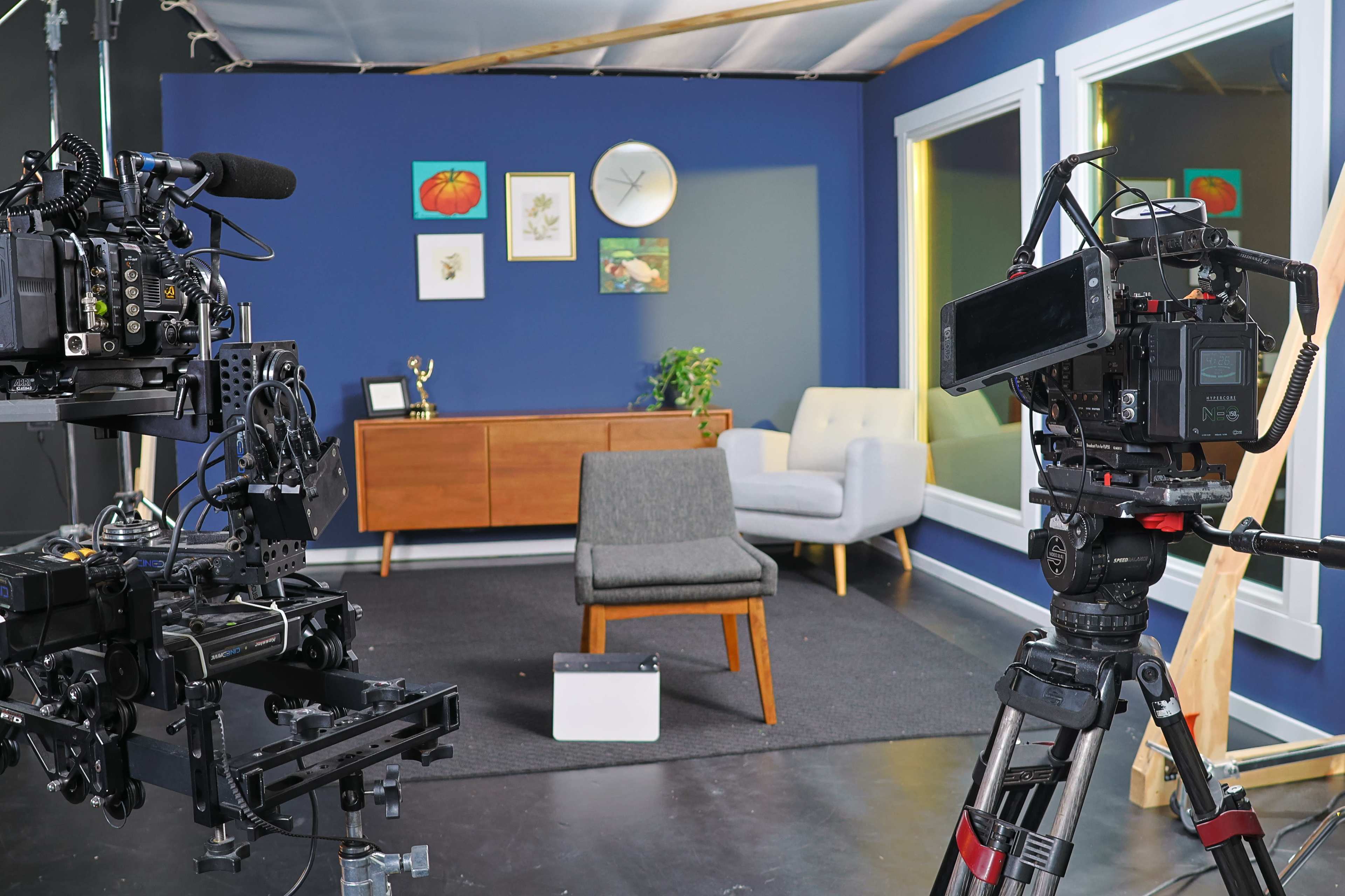A video recording setup featuring two cameras pointed at a minimalist studio space with a gray chair, a wooden desk, framed artwork, and large windows.