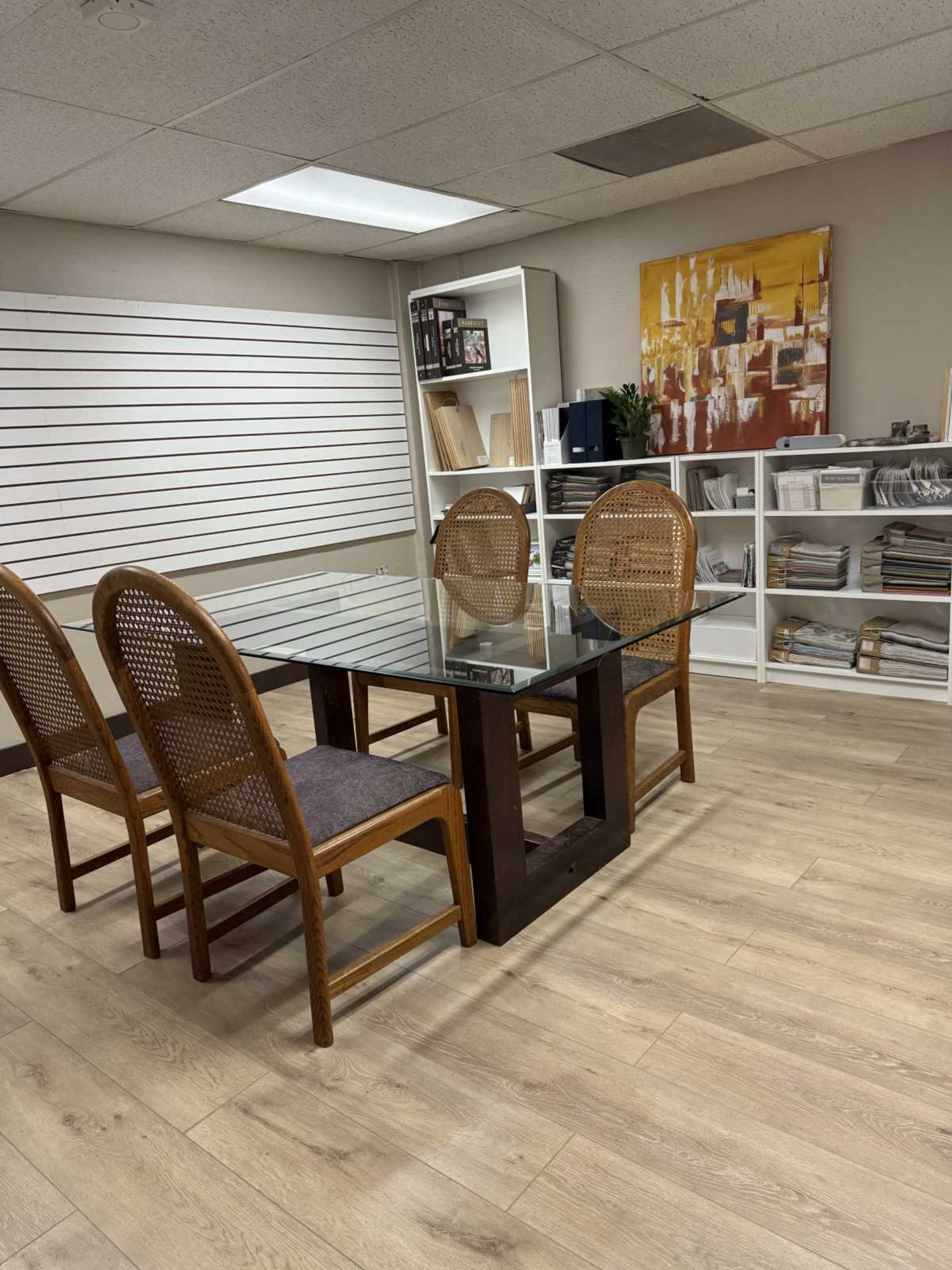 A glass-top table surrounded by four wooden chairs is situated in an office space with shelves displaying books and files.