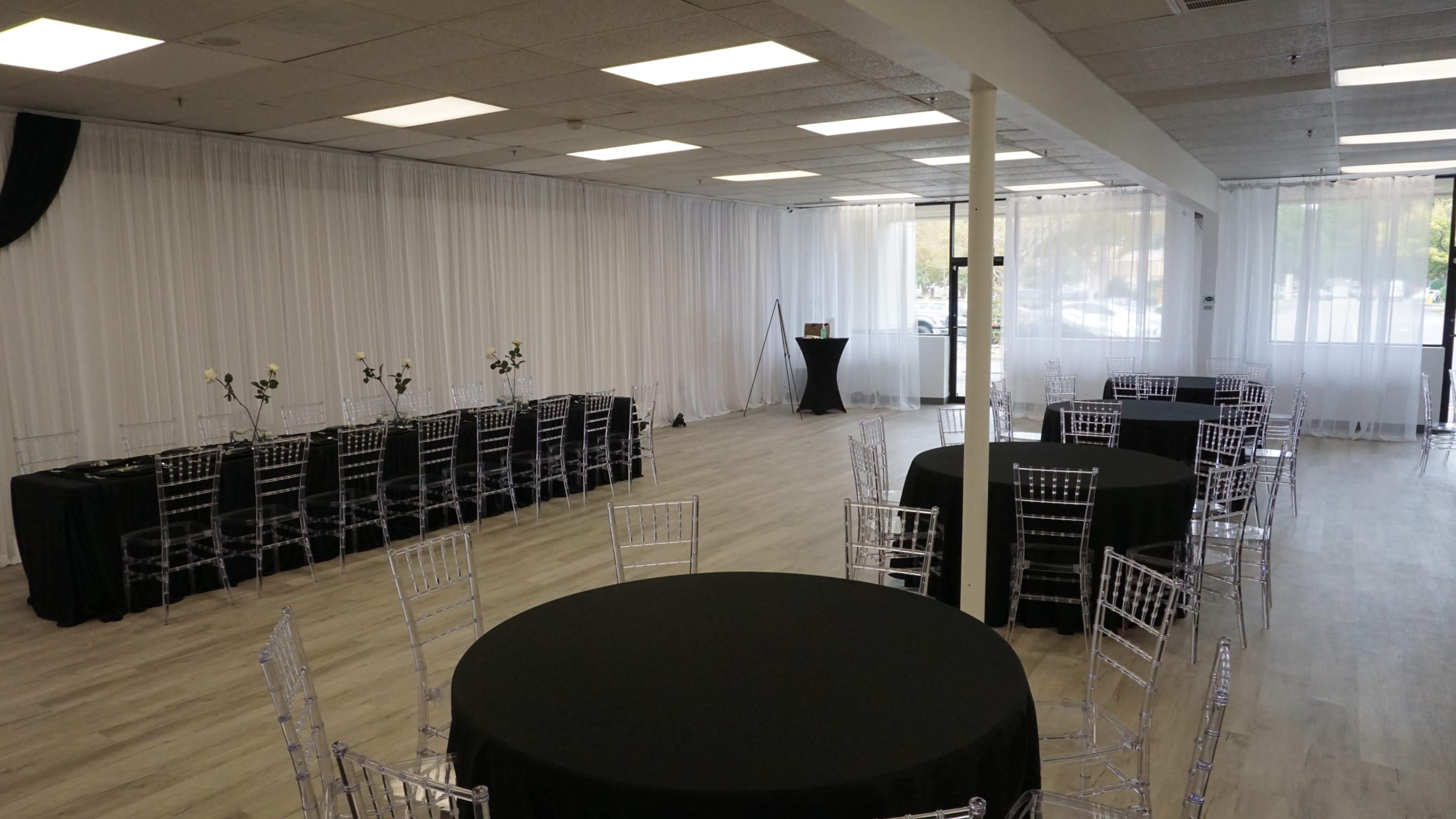The image shows an empty event space with black round tables and transparent chairs arranged in a clean, modern layout, surrounded by white drapes and bright overhead lighting.
