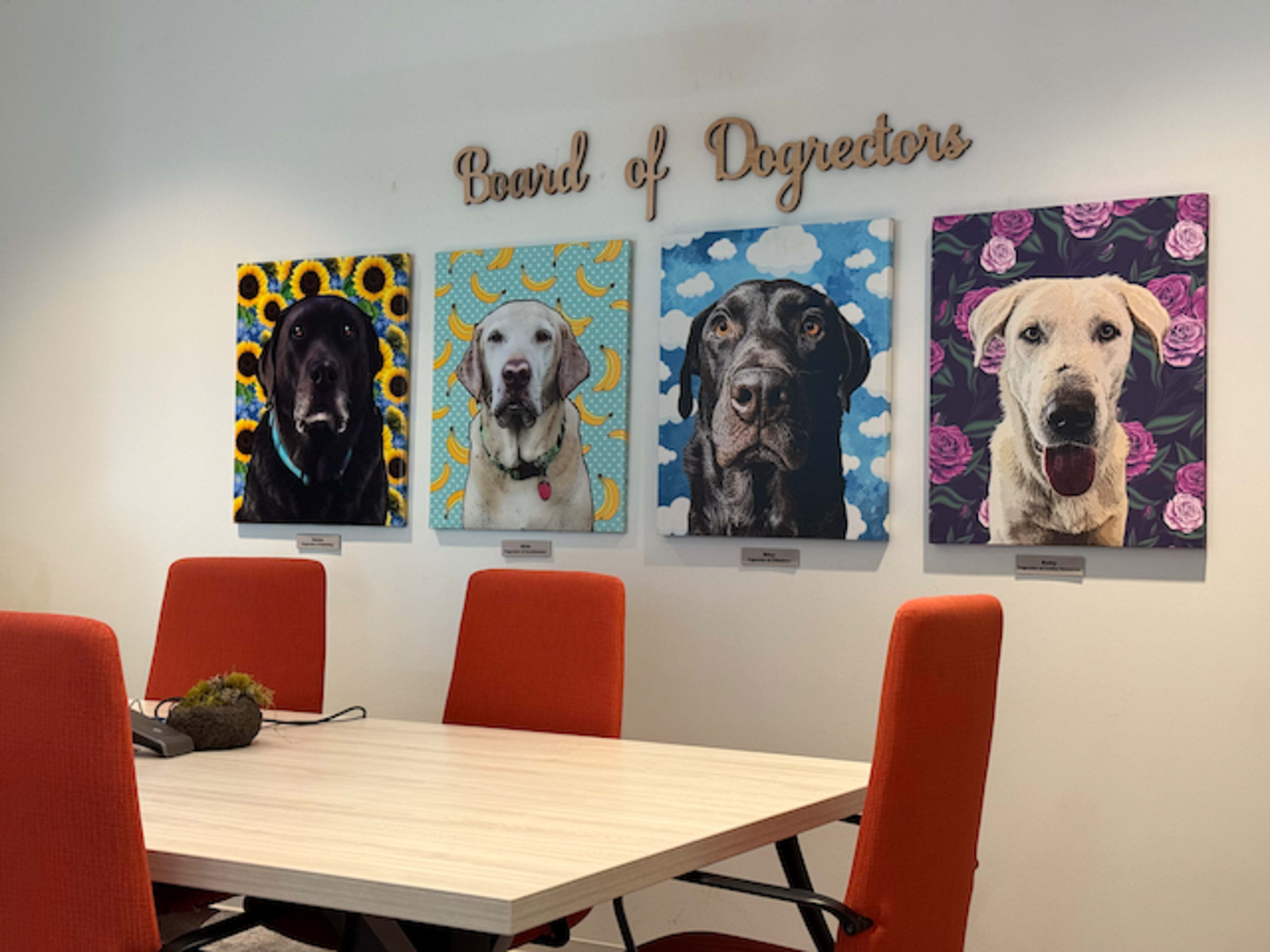 The image shows a conference room featuring four colorful dog portraits on the wall above a light wooden table and orange chairs.