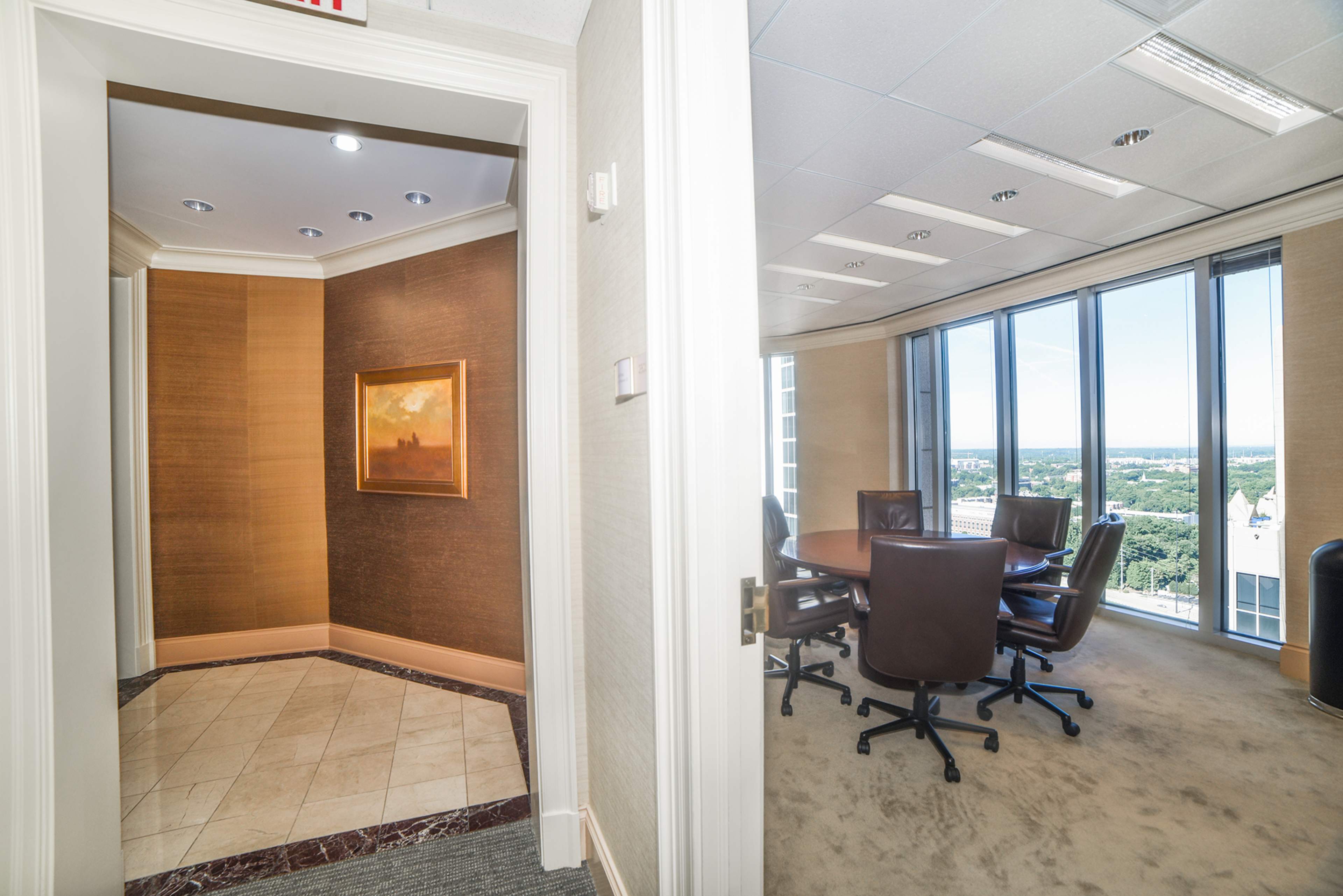 A well-lit office space with a conference table and chairs, visible through an entrance from a hallway featuring a painting on the wall.