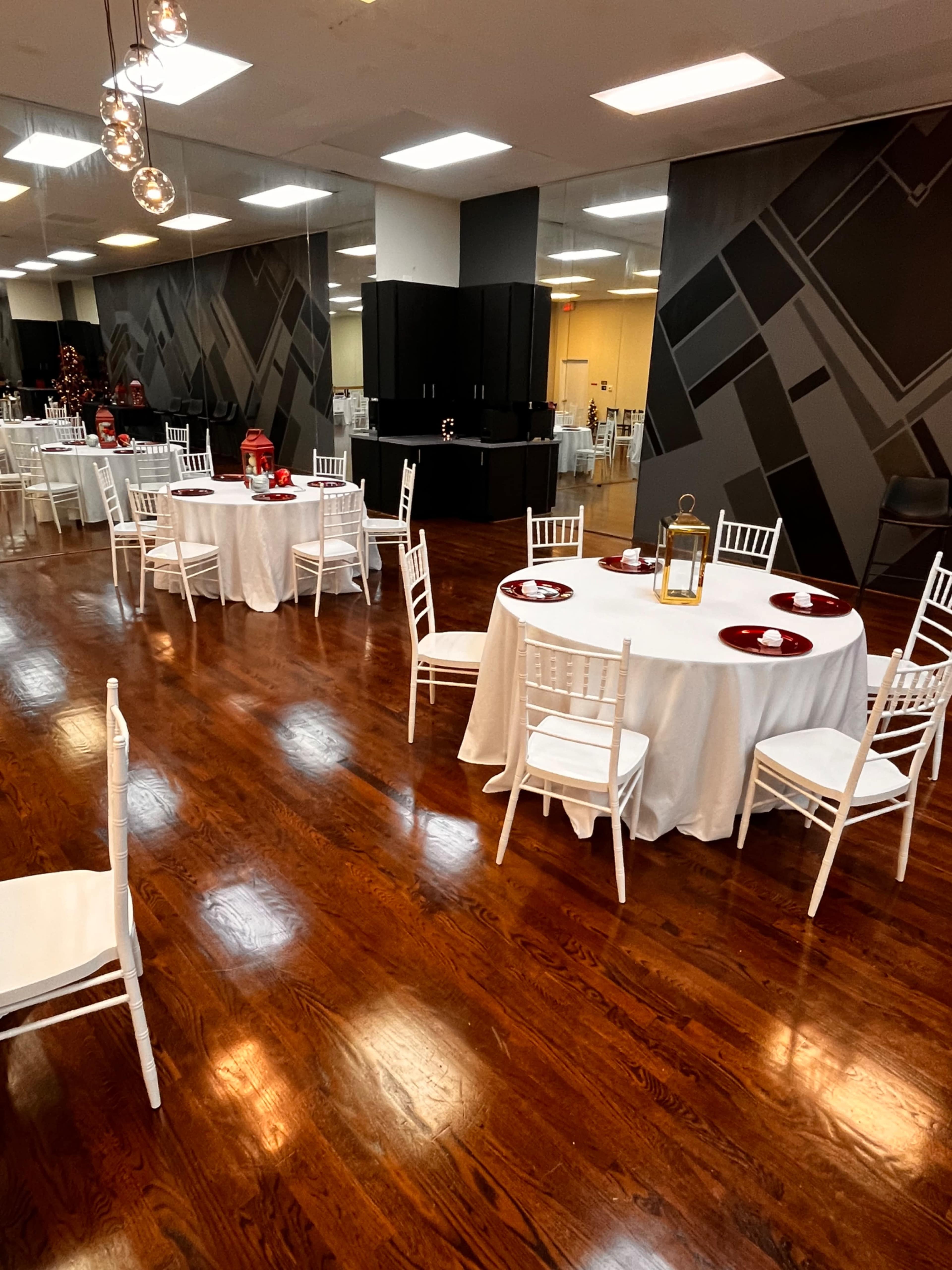 The image shows an elegantly arranged dining room with round tables covered in white tablecloths, each featuring red plates and surrounded by white chairs on a polished wooden floor.