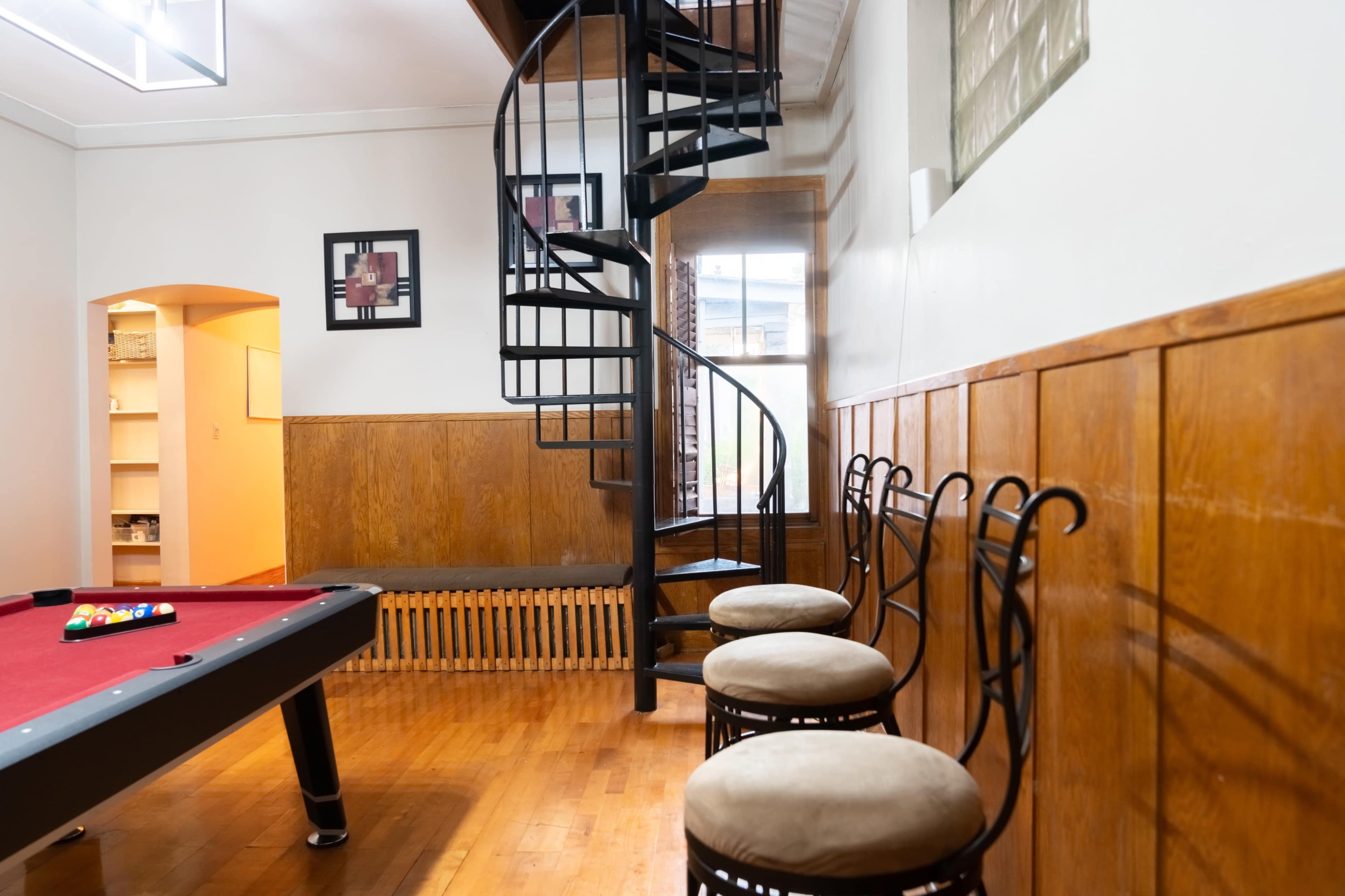 A spiral staircase leads to an upper level in a room featuring a pool table and several stools against a wooden wall.