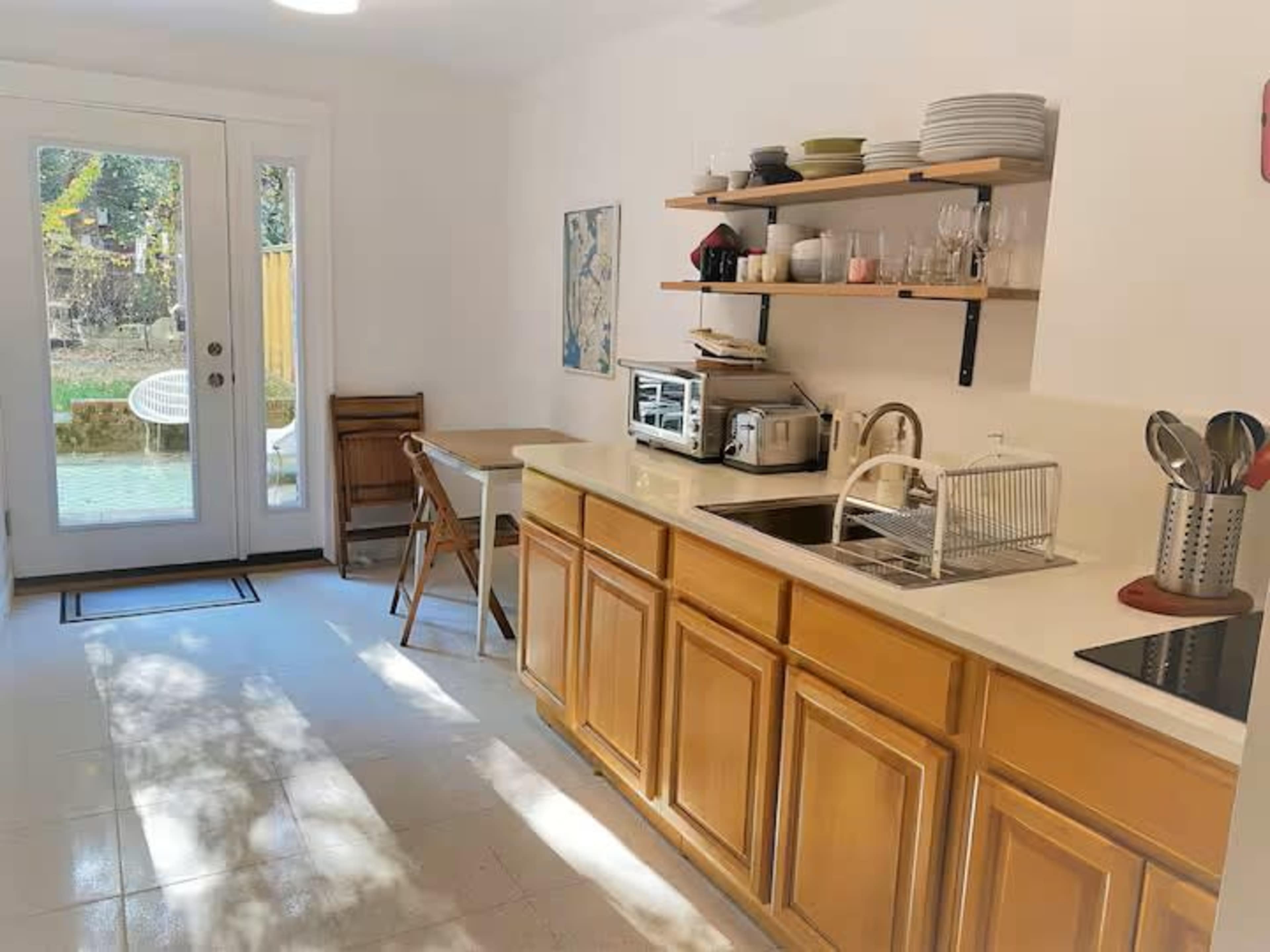 The image shows a modern kitchen with wooden cabinetry, a sink, various kitchen appliances, and dining furniture, alongside a door leading to an outdoor space.