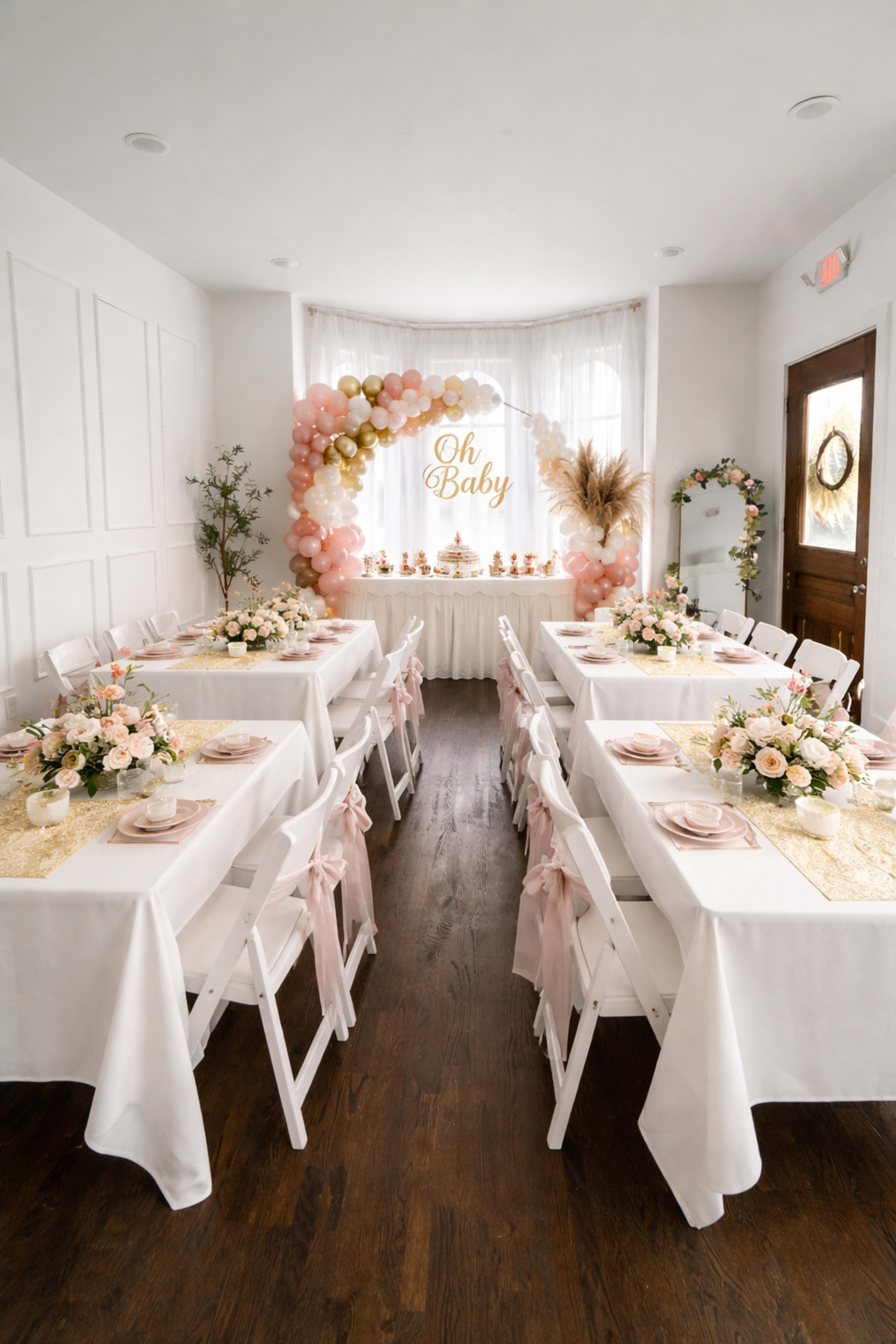 A decorated room features tables set for a baby shower, adorned with floral centerpieces and a balloon arch in the background.