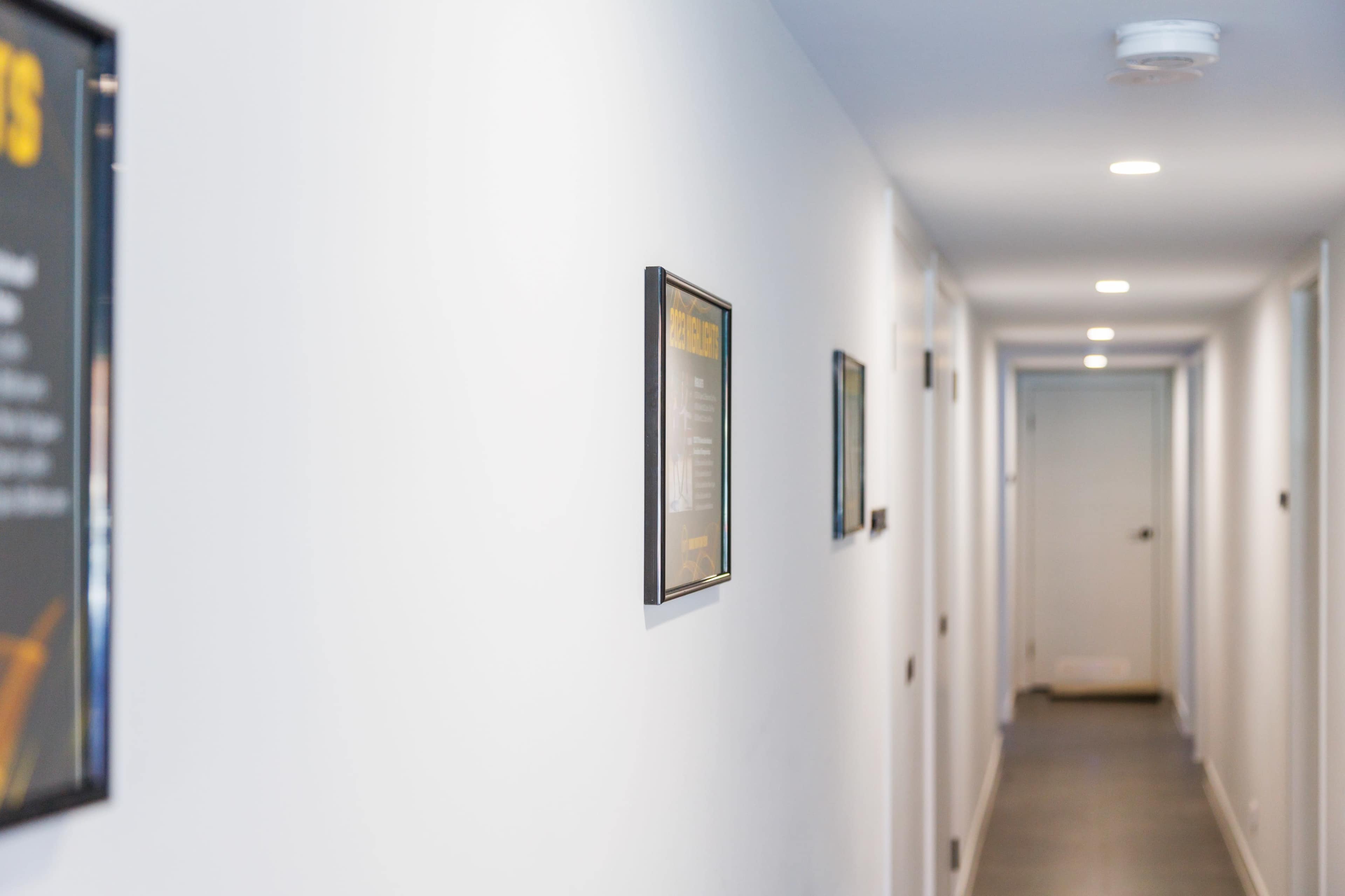 The image shows a narrow hallway with white walls, illuminated by ceiling lights, and framed pictures hung on one side.