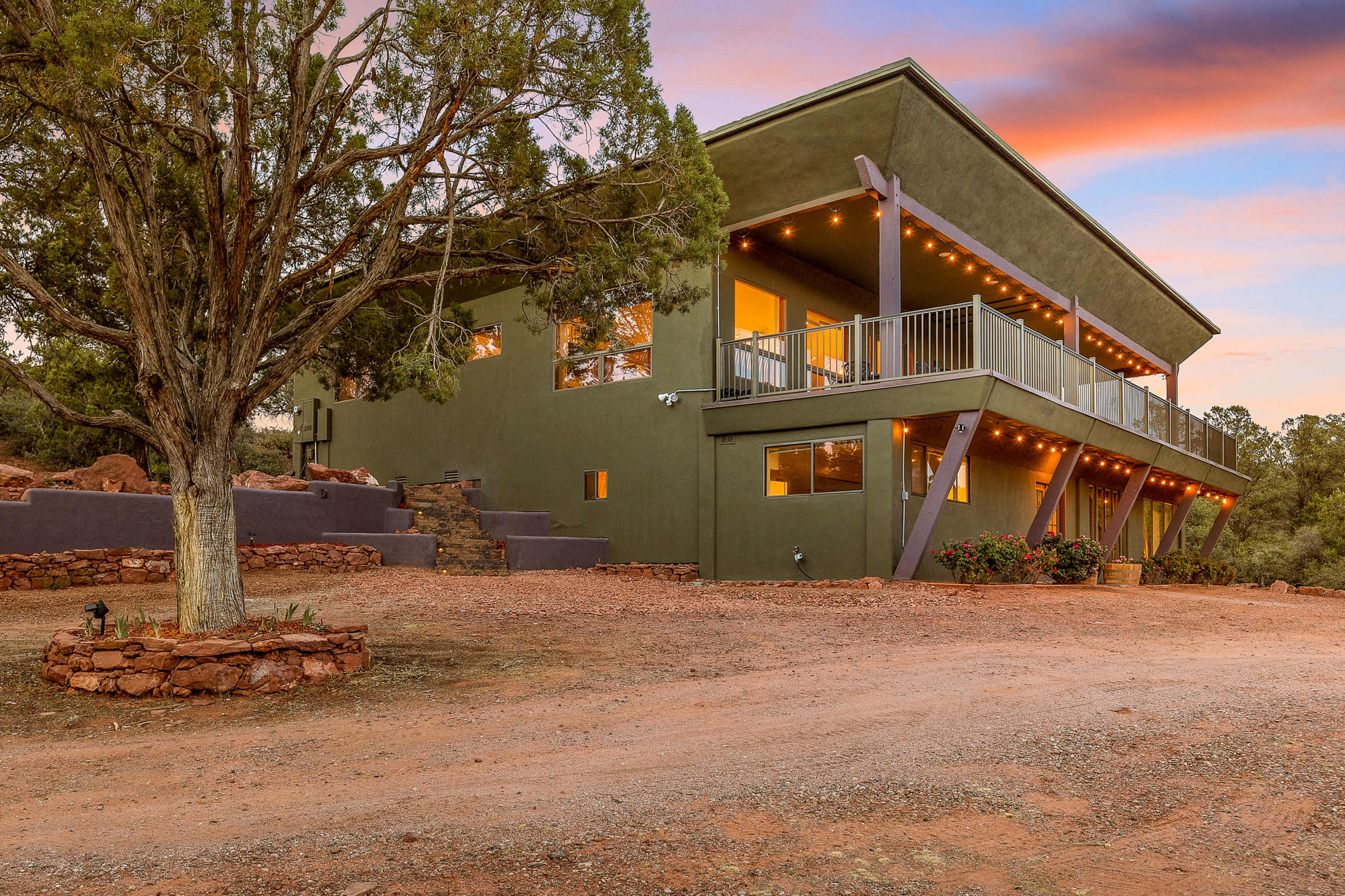 A modern two-story green house with a wraparound deck and string lights is situated on a gravel driveway surrounded by trees.