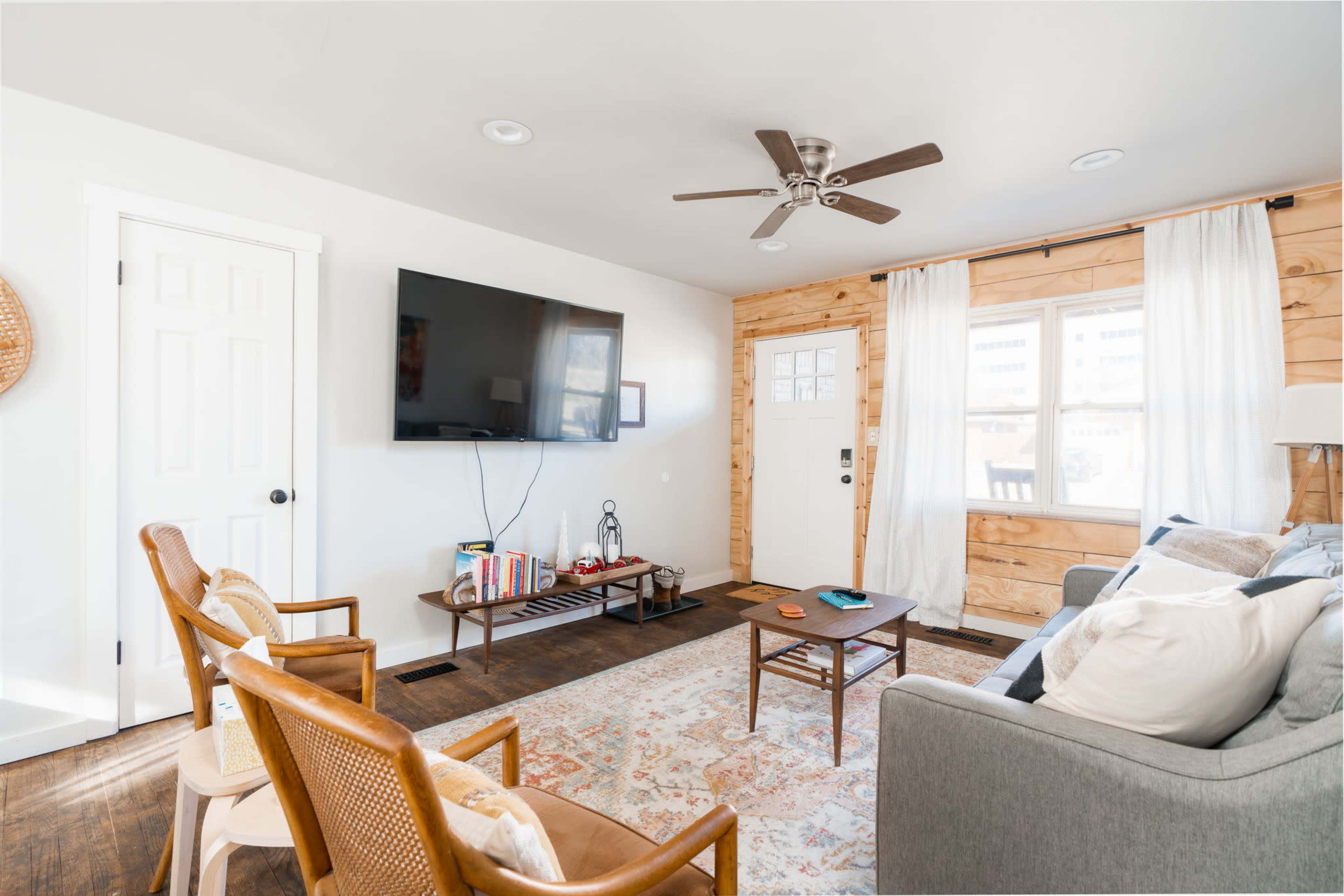 The image shows a living room with a gray sofa, a coffee table, and a television mounted on the wall, featuring wooden accents and large windows with sheer curtains.