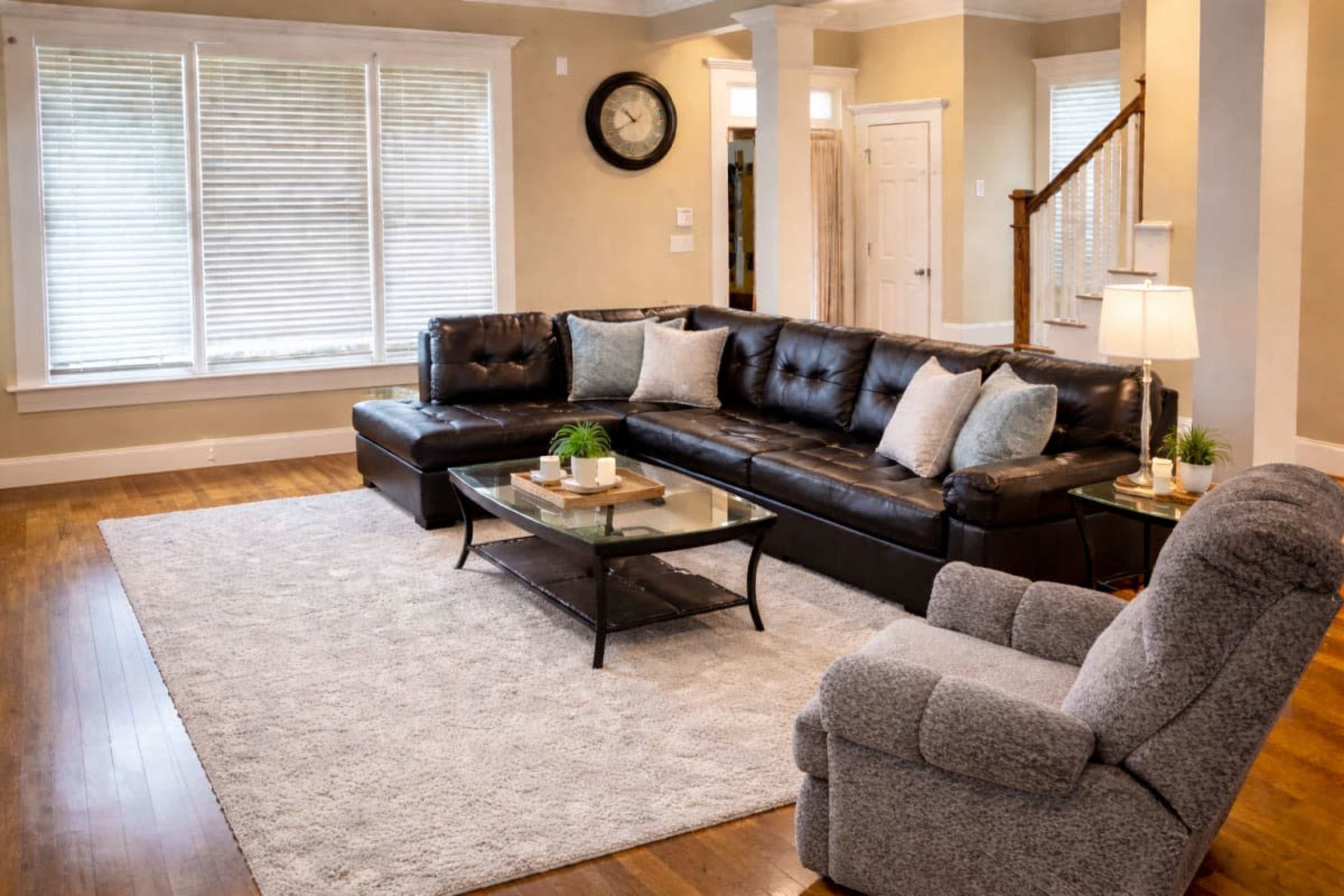 The image features a spacious living room with a dark leather sectional sofa, a glass coffee table, and a cozy armchair on a light-colored area rug.