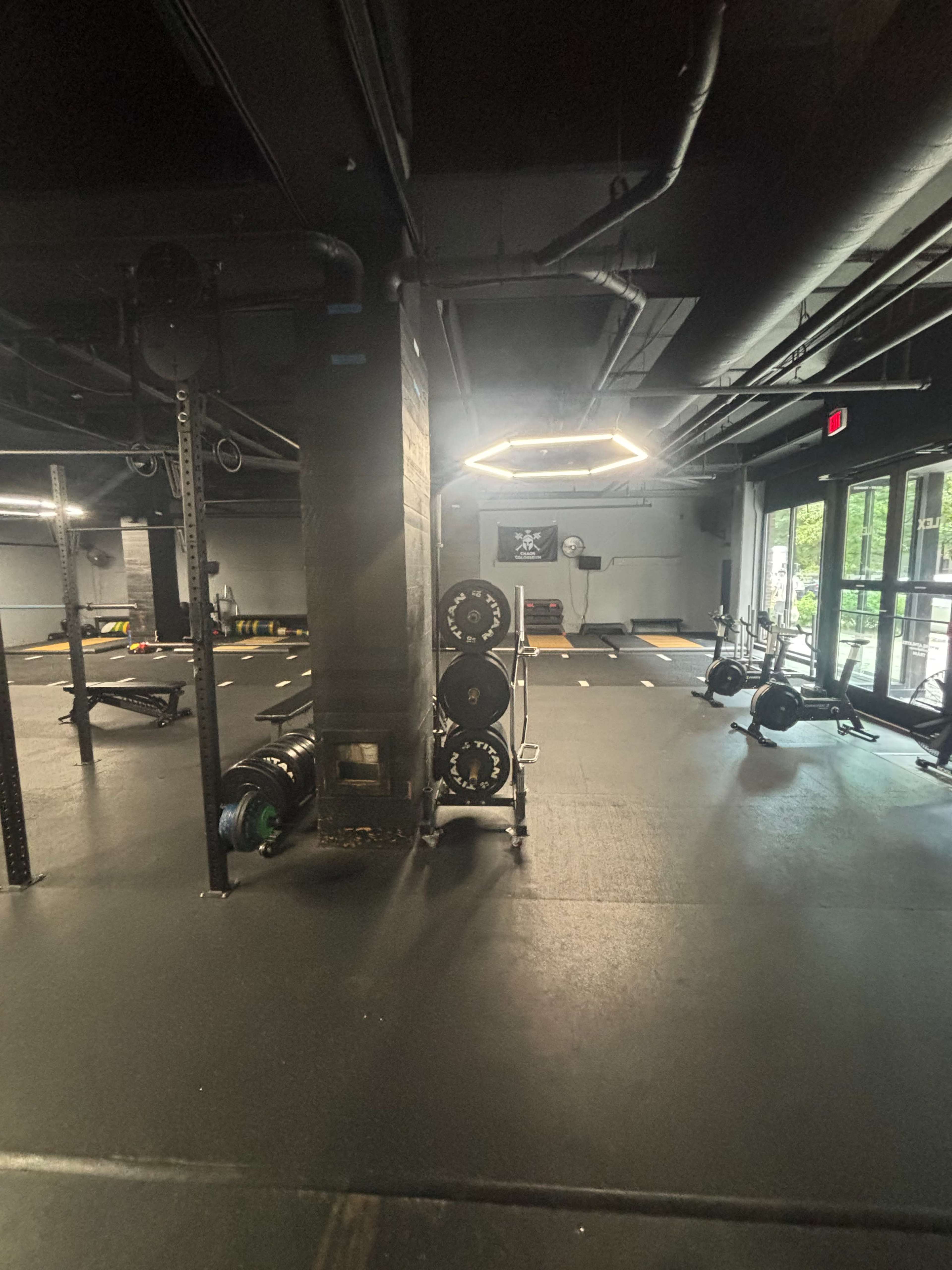 The image shows an empty fitness gym with workout equipment arranged on black flooring and large windows letting in natural light.