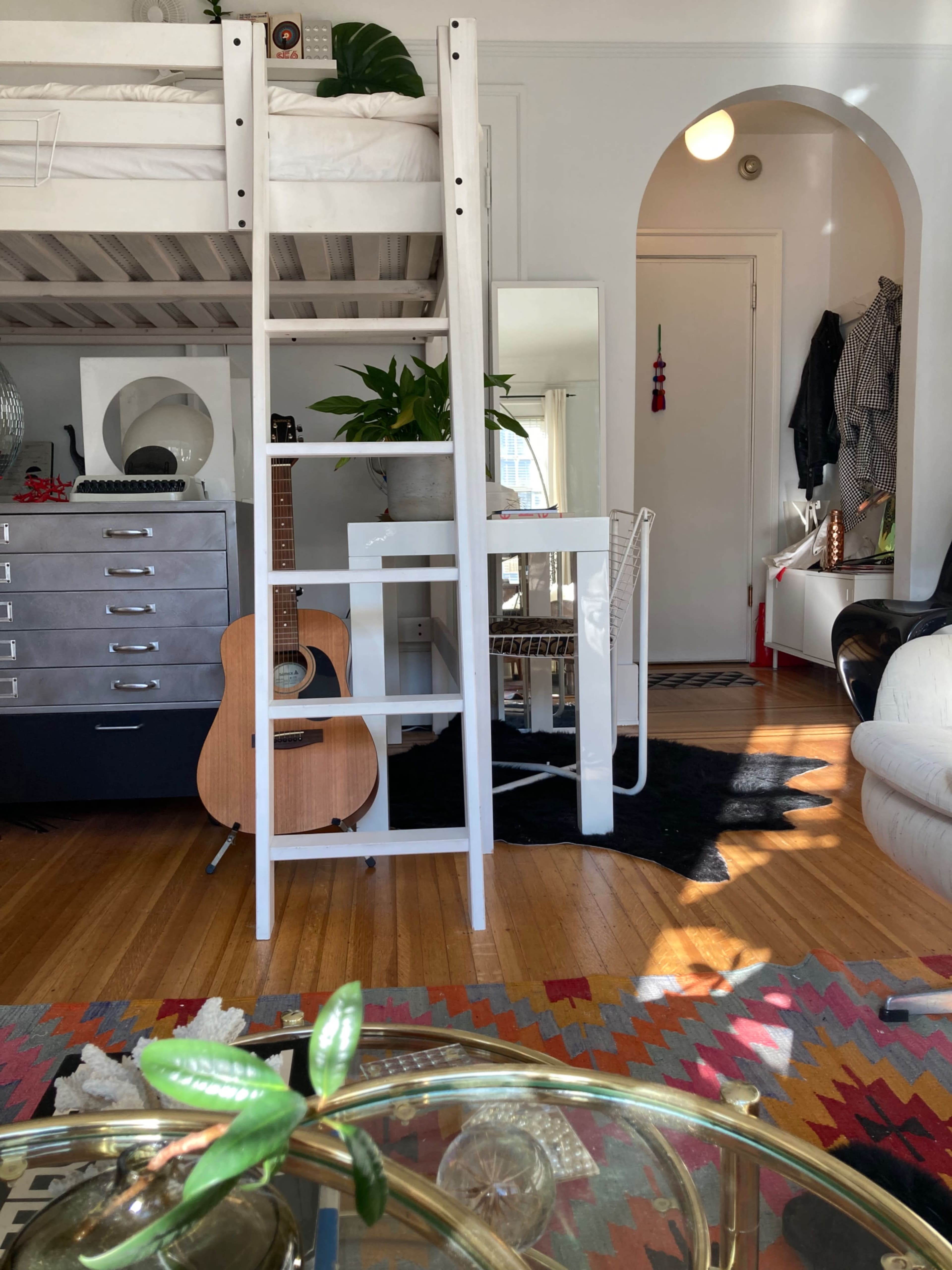 A lofted bed is positioned above a white table and chair, with an acoustic guitar leaning against a dresser in a brightly lit room featuring wooden floors and a colorful rug.