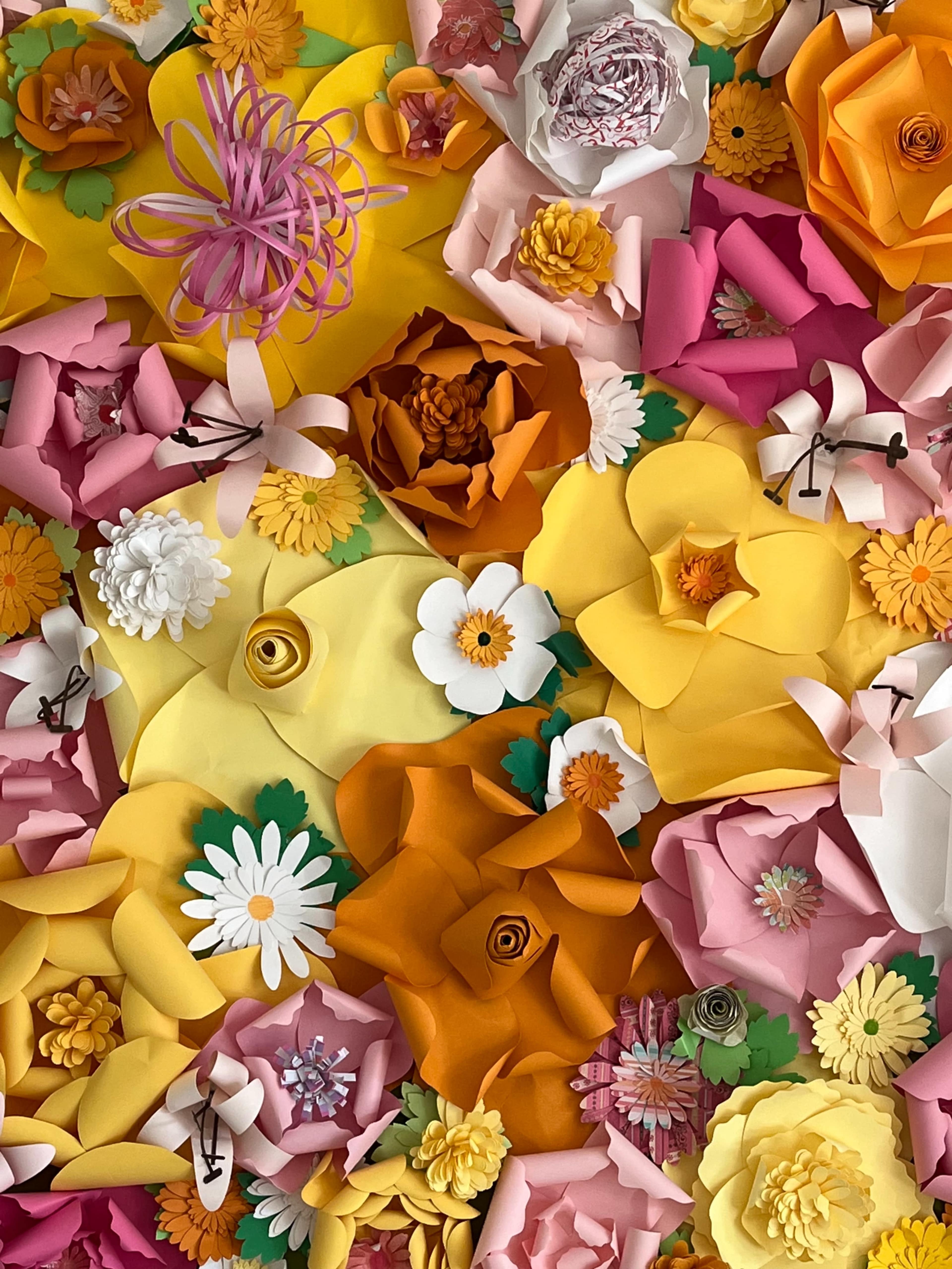 The image displays a vibrant arrangement of various paper flowers in shades of yellow, orange, pink, and white, set against a colorful backdrop.