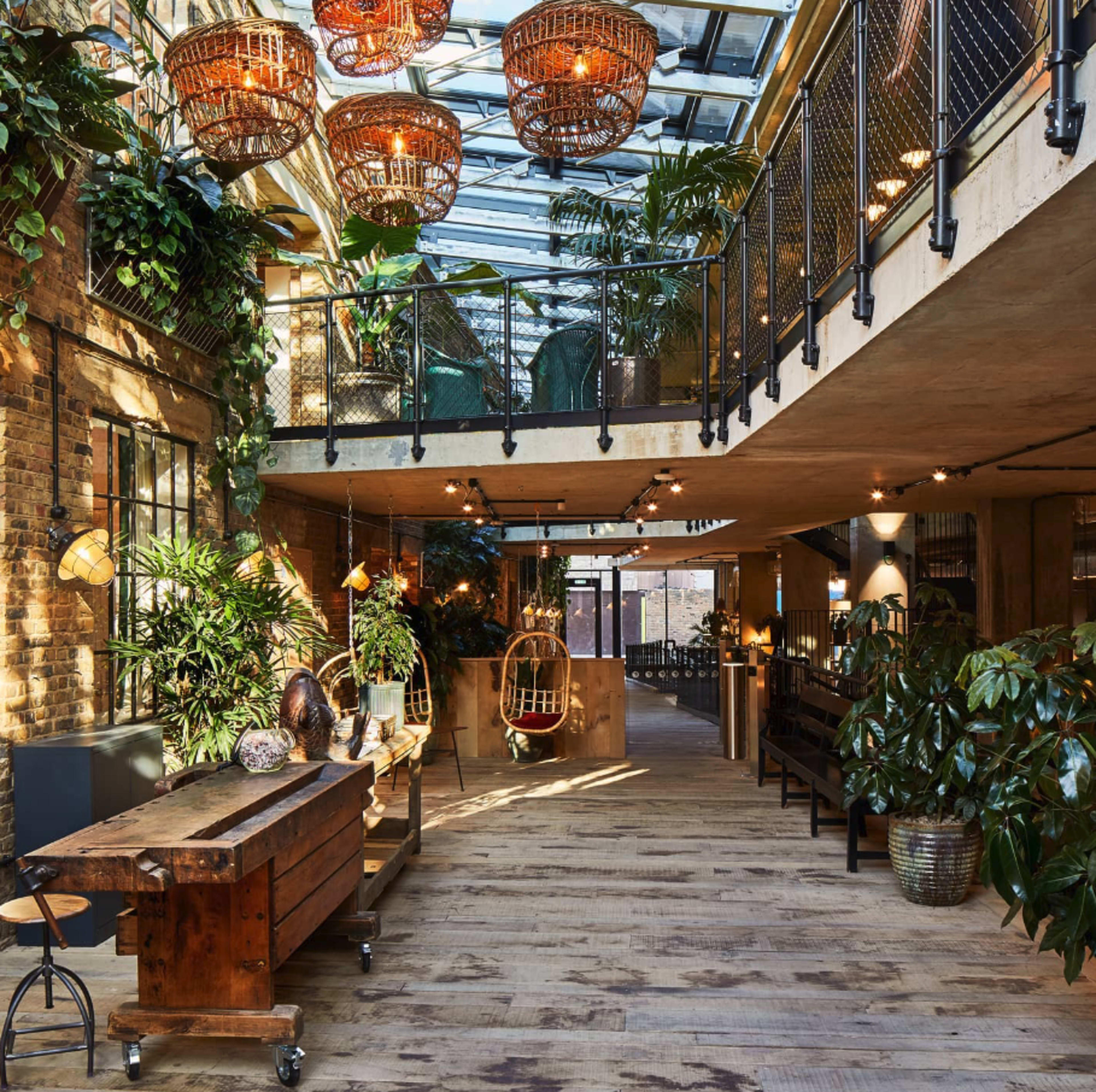 The image shows a modern interior space featuring a wooden table, hanging light fixtures, and abundant greenery under a glass ceiling.